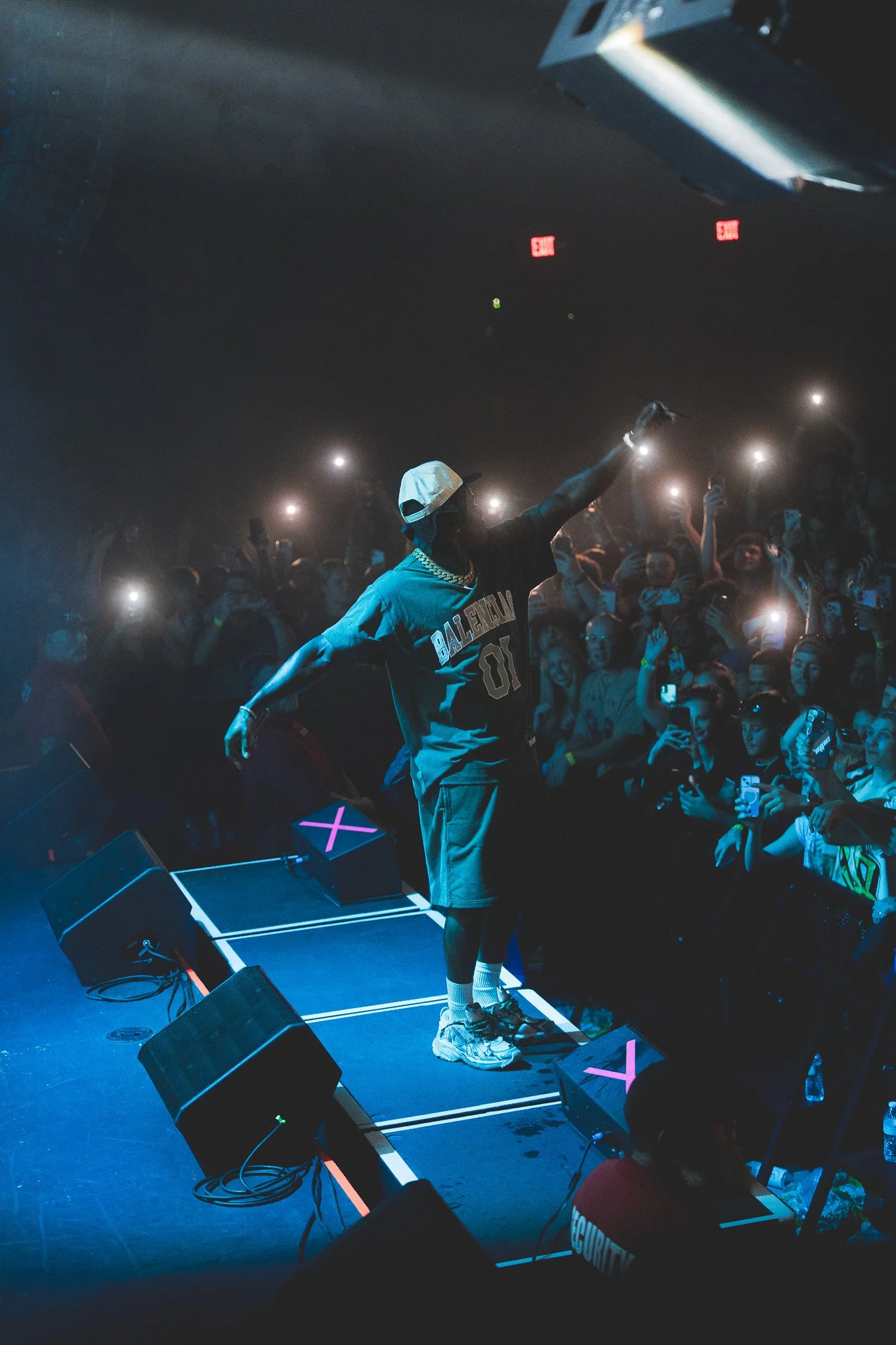 DaBaby on stage at a concert, wearing a Balenciaga jersey, shorts, sneakers, and a white cap, with one arm extended and surrounded by a crowd of fans taking photos and videos.