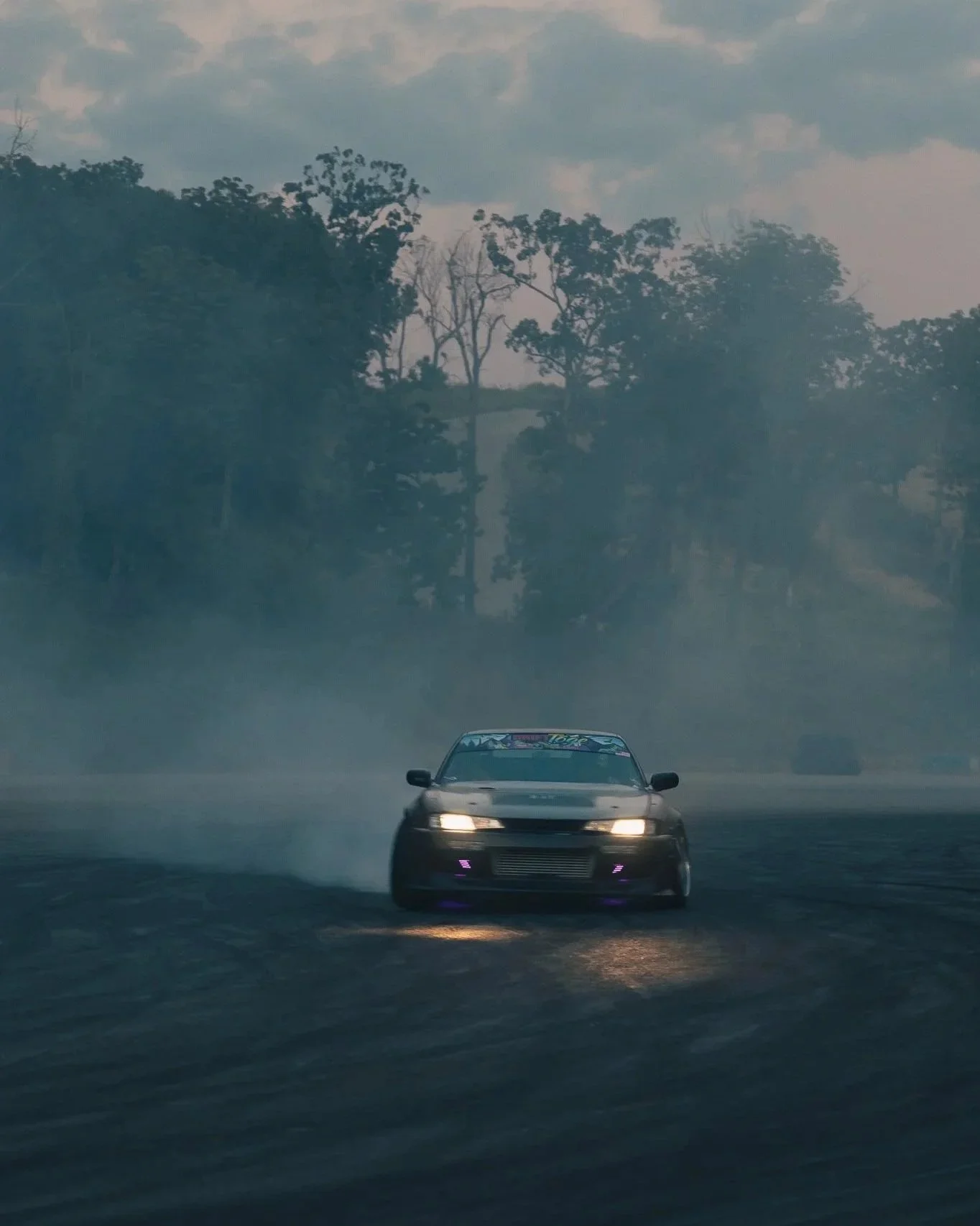 A black sports car drifting on a wet race track with water spray and mist, surrounded by trees and an overcast sky.