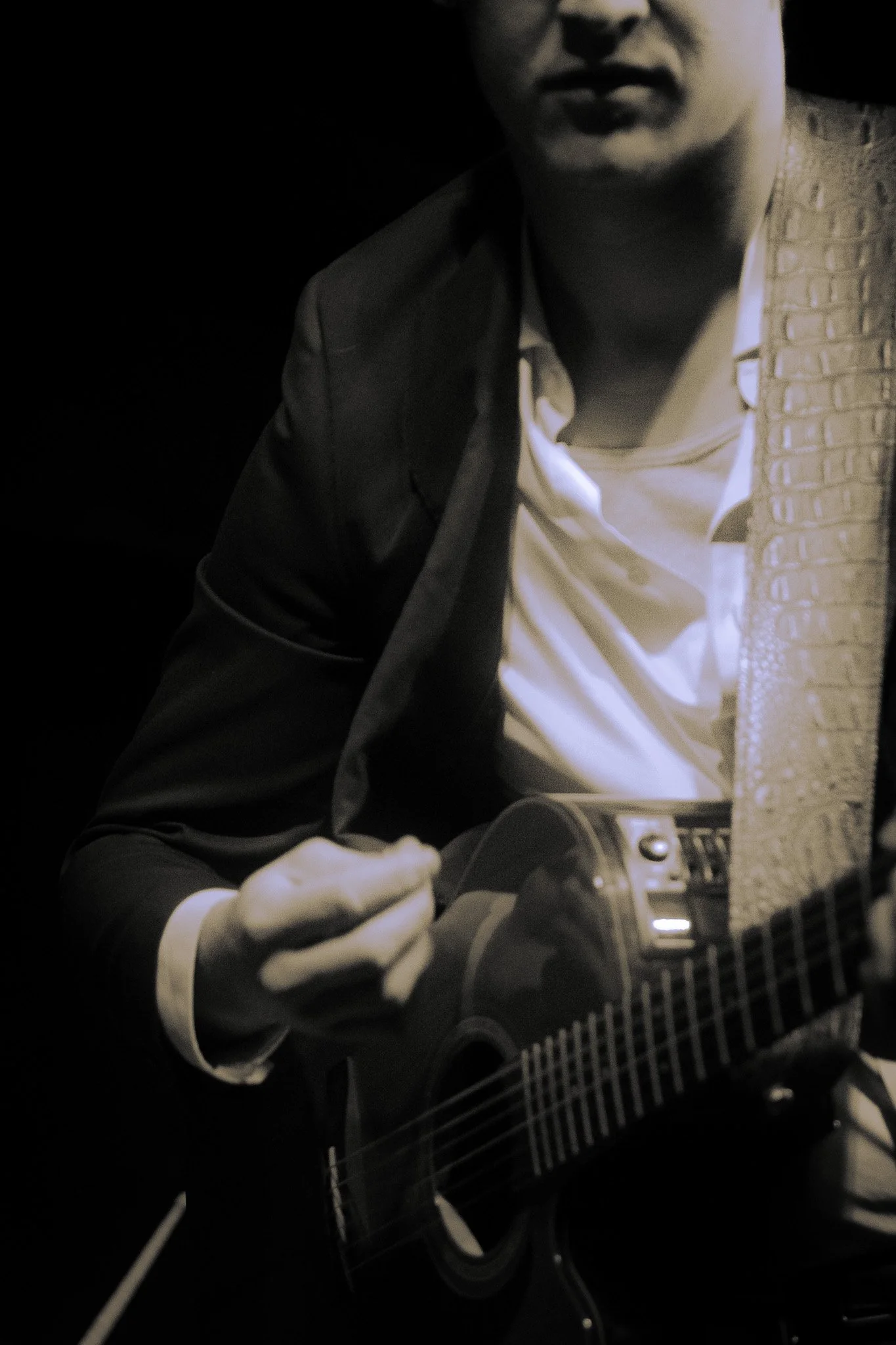 A man wearing a suit jacket and white shirt, playing an acoustic guitar in a dark setting.
