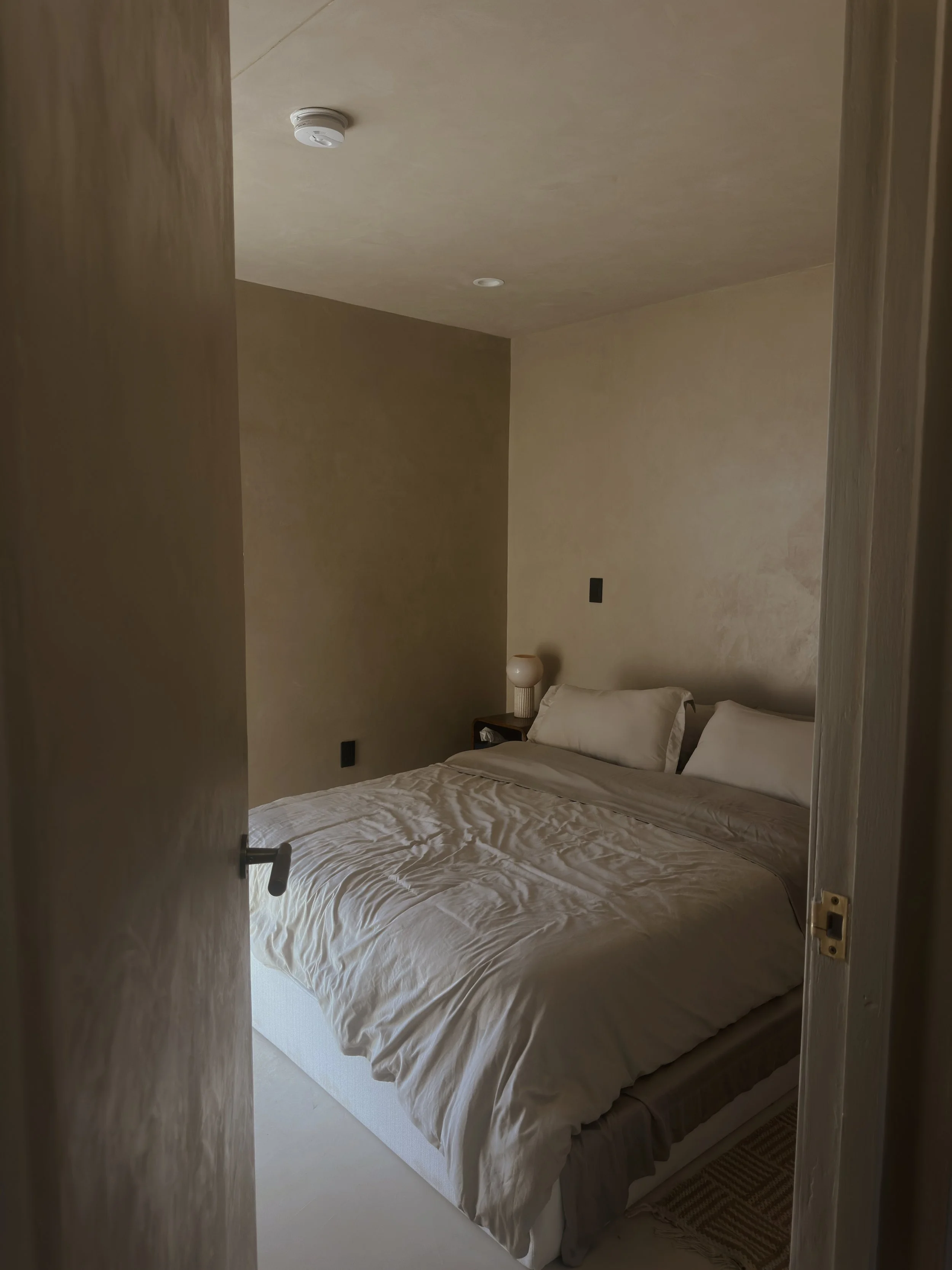 A neatly made bed with white bedding and two pillows, positioned against a beige wall in a minimalistic bedroom. There is a dark-colored nightstand with a white lamp next to the bed, and the door to the room is partially open.
