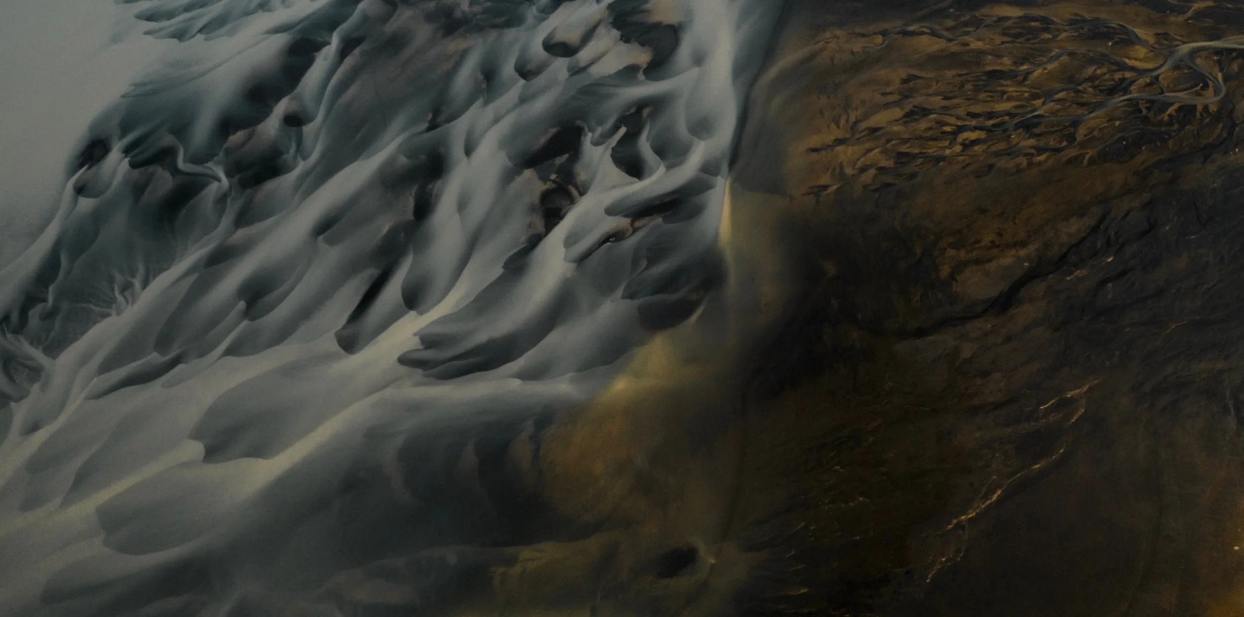 Aerial view of a glacier with dark, rugged terrain and a blue ice formation with crevasses.
