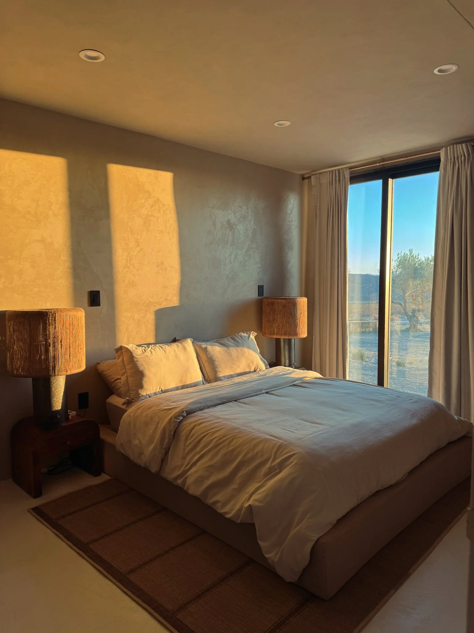 A bedroom with a bed, two bedside tables with lamps, and large windows with curtains letting in sunlight.