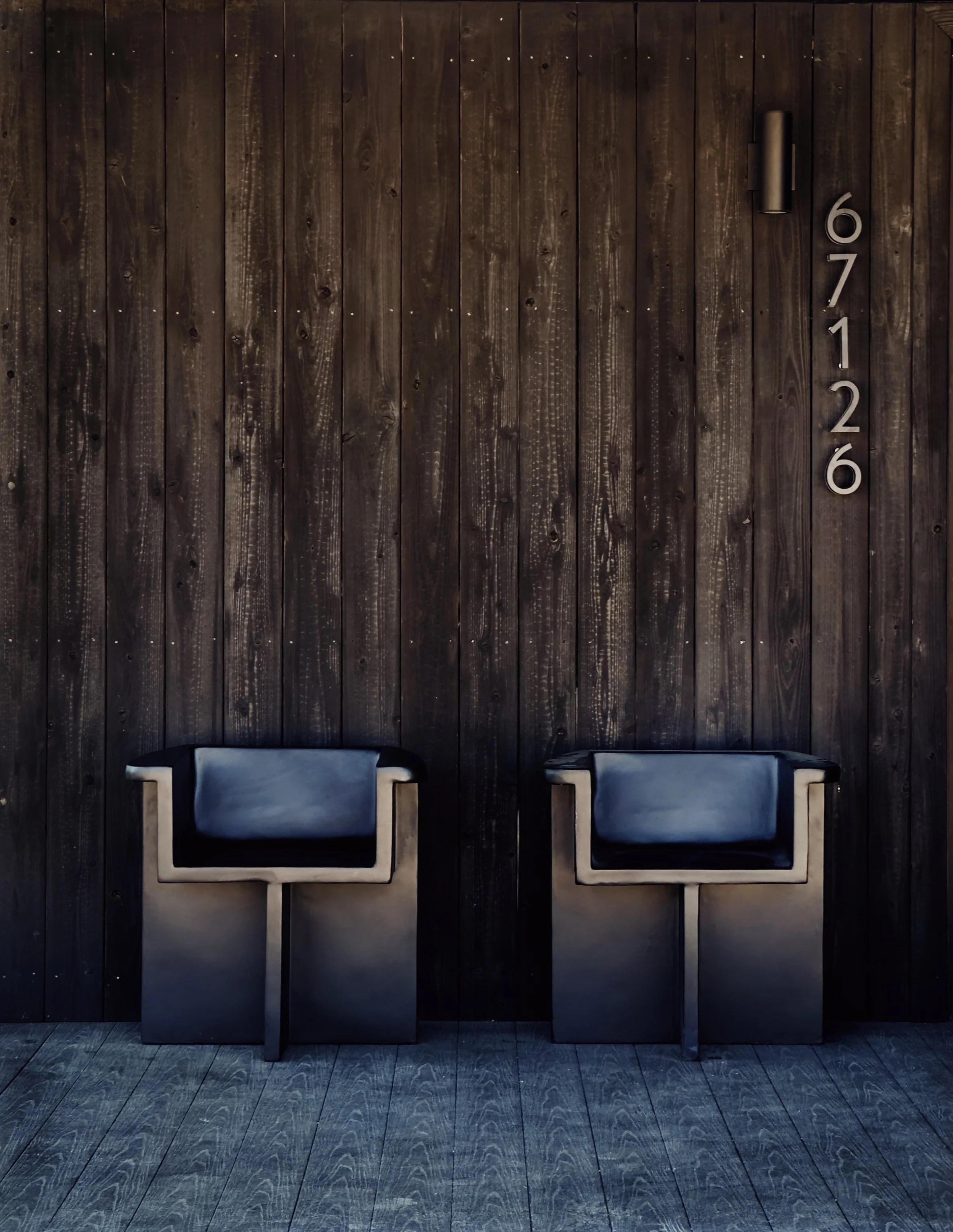 Two modern black armchairs with wooden accents are placed against a dark wooden wall, with a metallic house number 671226 vertically aligned on the right side of the wall.