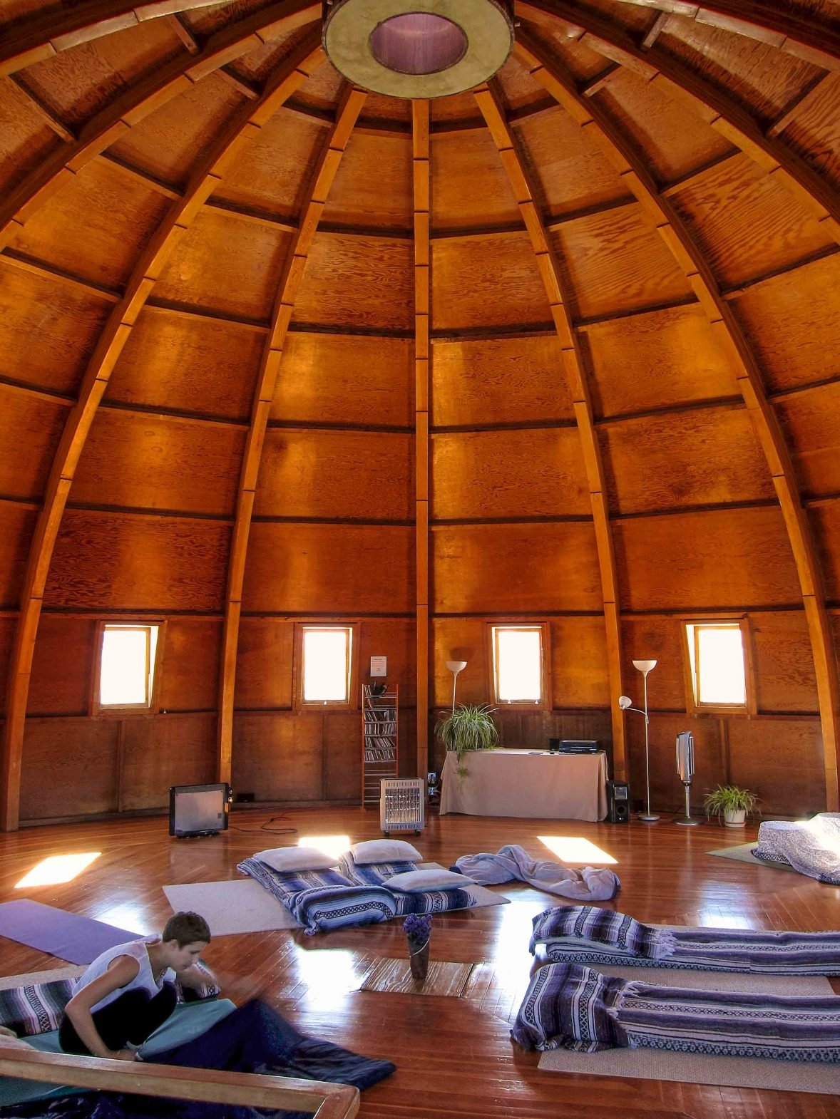 Interior of a wooden dome with four small windows, a person sitting on cushions, and various plants, lamps, and speakers in a spacious, circle-shaped room.
