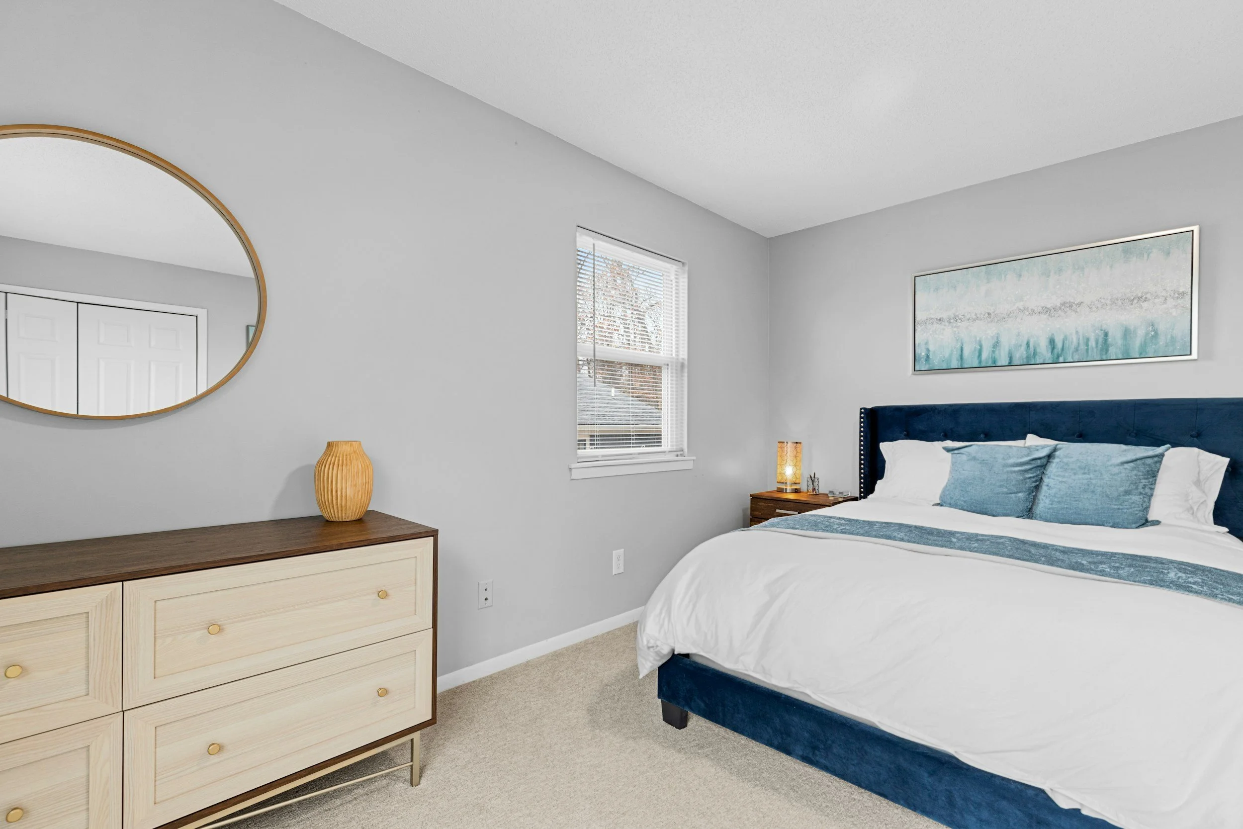 Bedroom with light gray walls, a large bed with a navy blue headboard, white and light blue bedding, a wooden dresser with a round mirror and a yellow vase, a small nightstand with a lamp and decorative items, and a framed painting above the bed.