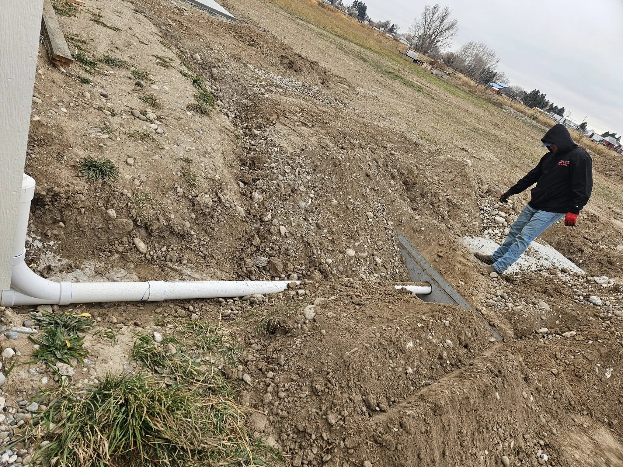 A person wearing a black hoodie, blue jeans, and red gloves inspecting a drainage pipe outside on a construction site with exposed dirt and gravel.