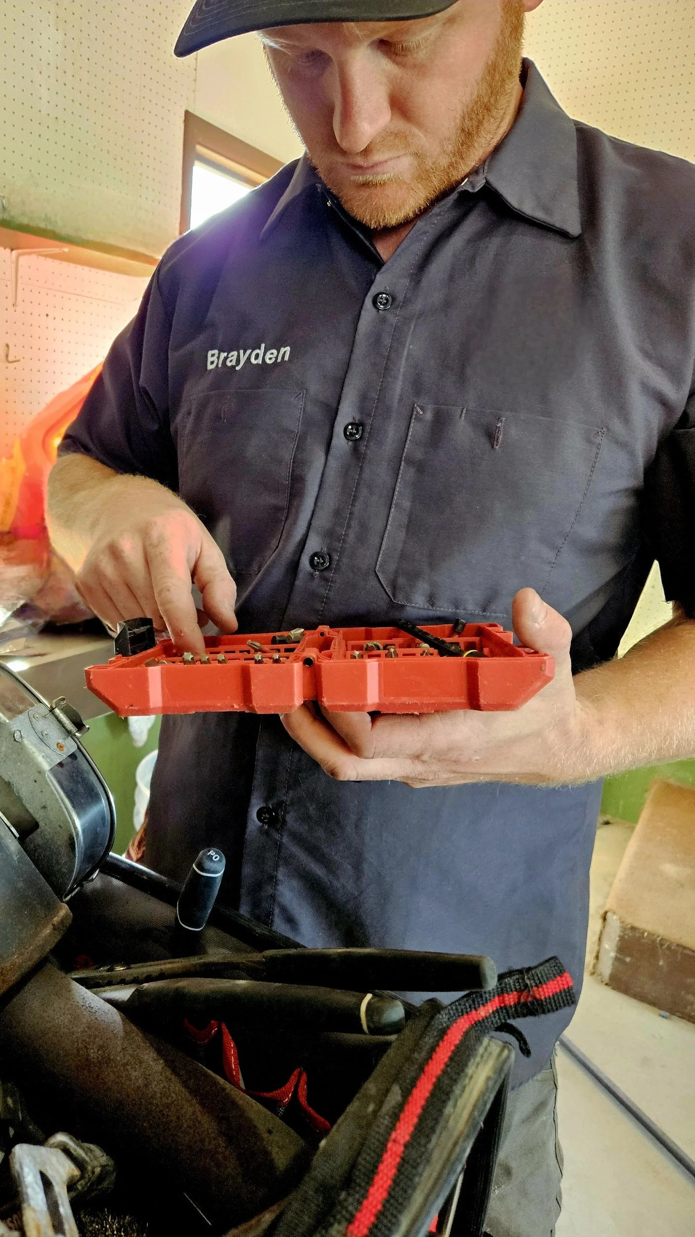 A man wearing a gray work shirt with the name 'Brayden' is looking down while holding a small red toolbox and using a tool on it.