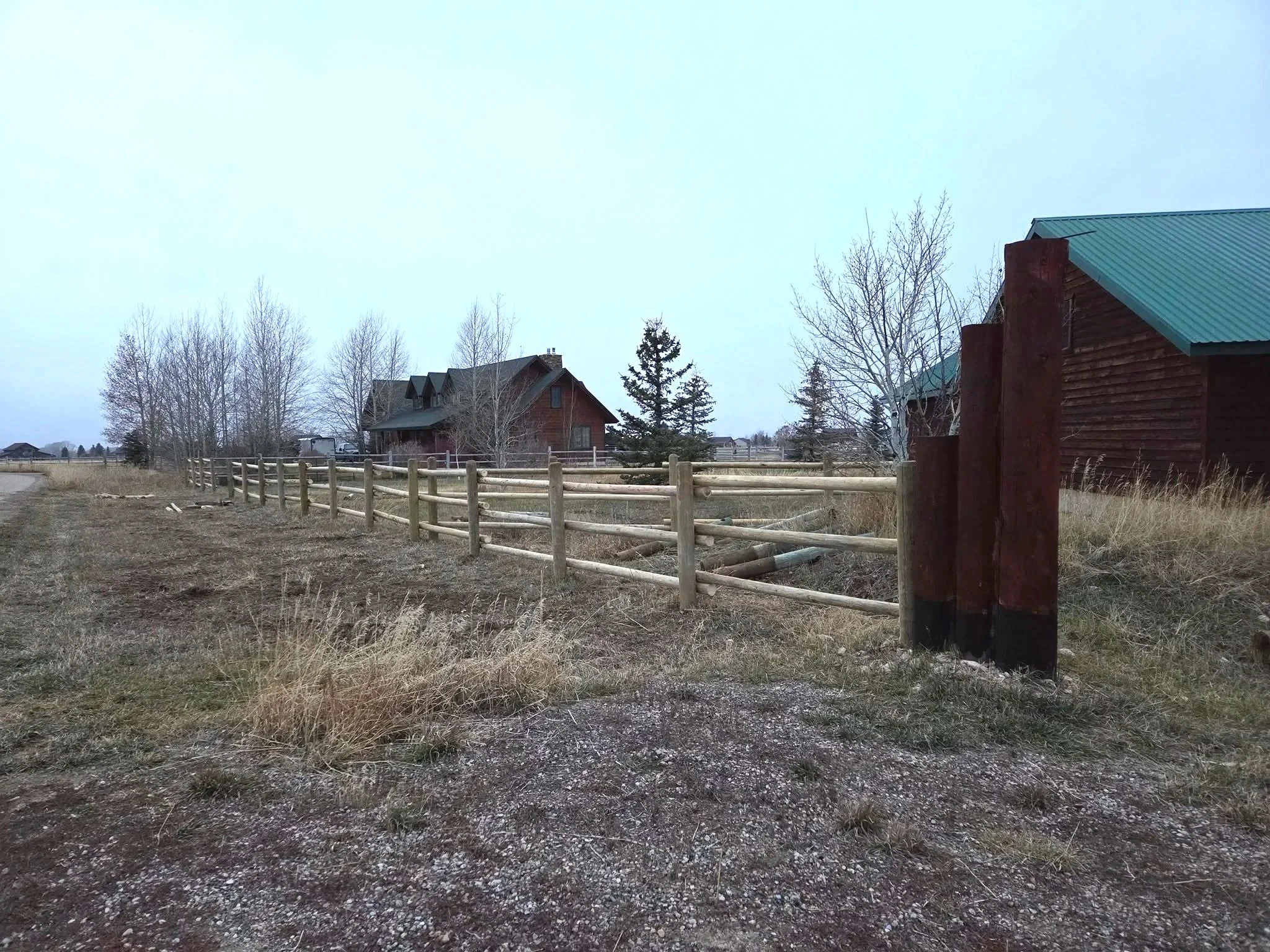 A wooden fence runs along a gravel path outside a rural property, with a house and some trees in the background under an overcast sky.