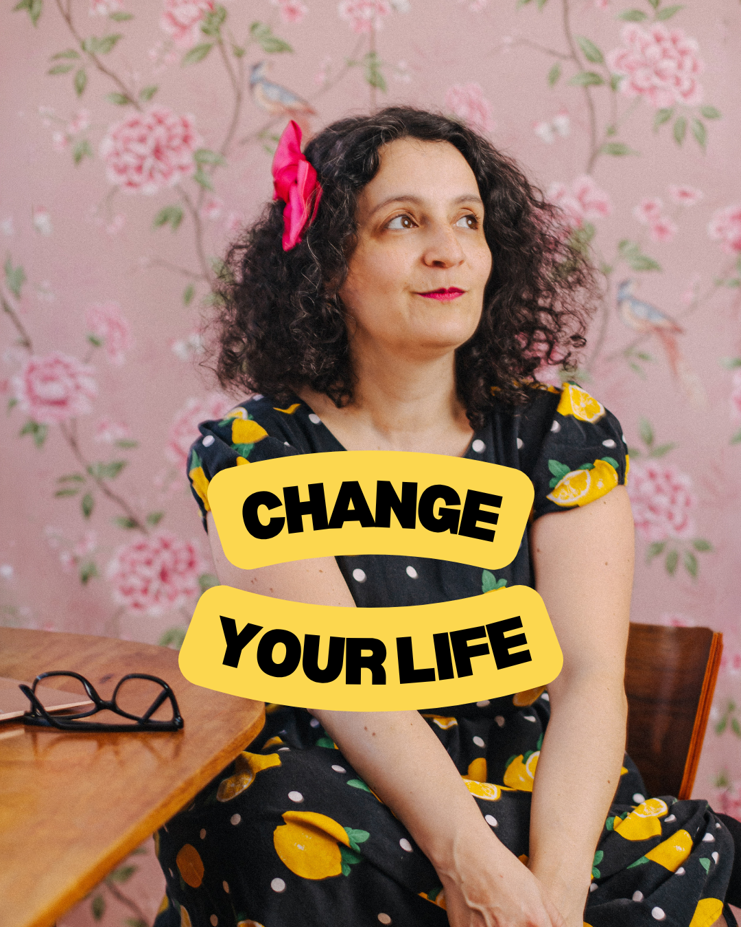 A woman with curly dark hair wearing a colorful dress with lemons and polka dots, sitting at a wooden table with glasses on it, against a pink floral wallpaper background, with a pink hair bow, and the text 'Change Your Life' in bold black letters on yellow banners.