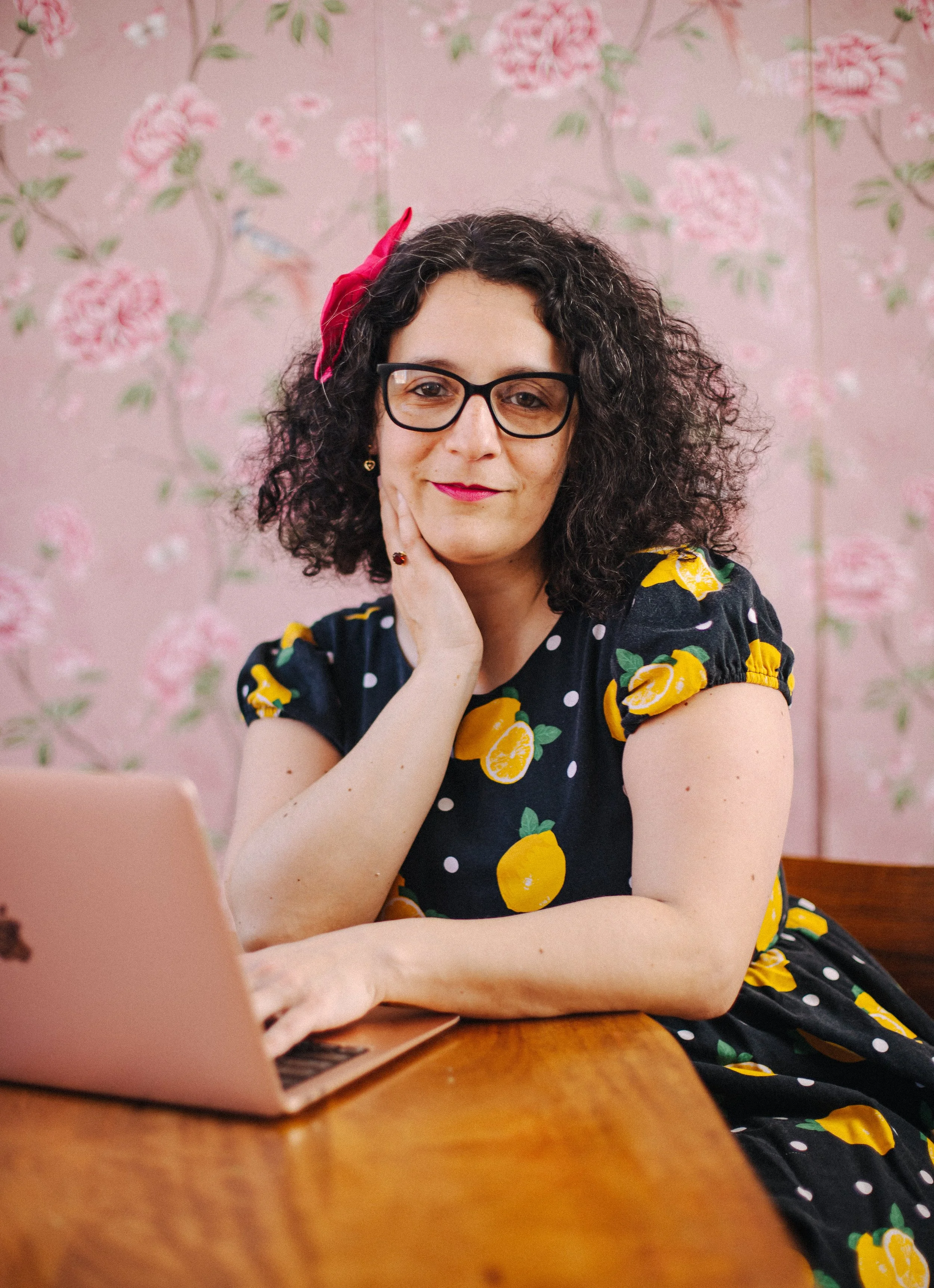A woman with curly dark hair, wearing glasses and a red hair bow, sitting at a wooden table with a pink laptop. She is wearing a black dress with a lemon print, and has her hand resting on her chin. The background is pink with floral patterns.