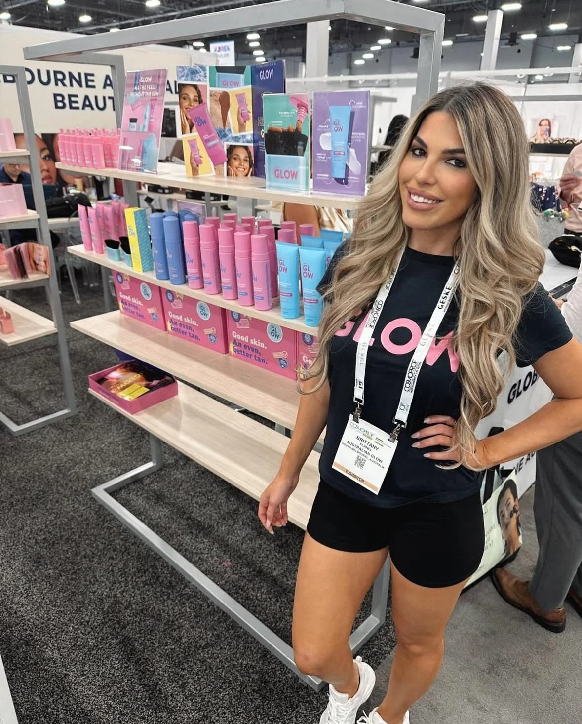 A woman with long blonde hair smiling and wearing a black t-shirt with pink letters and black shorts at a beauty product booth. The booth displays pink, blue, and purple skincare or cosmetic products on shelves.