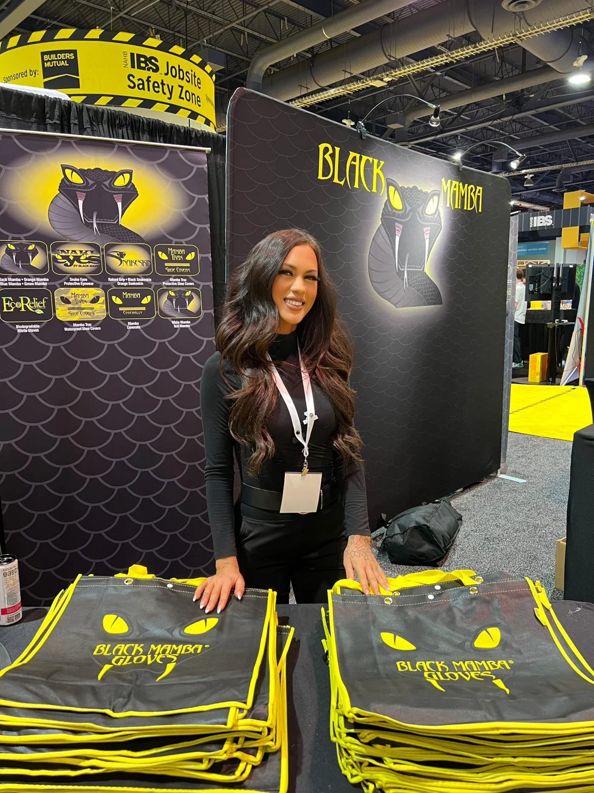 A woman standing behind a booth at a trade show, displaying yellow and black gloves with black mamba snake logo. The booth features black and yellow black mamba branding and product display on the table, with background banners showcasing the snake l