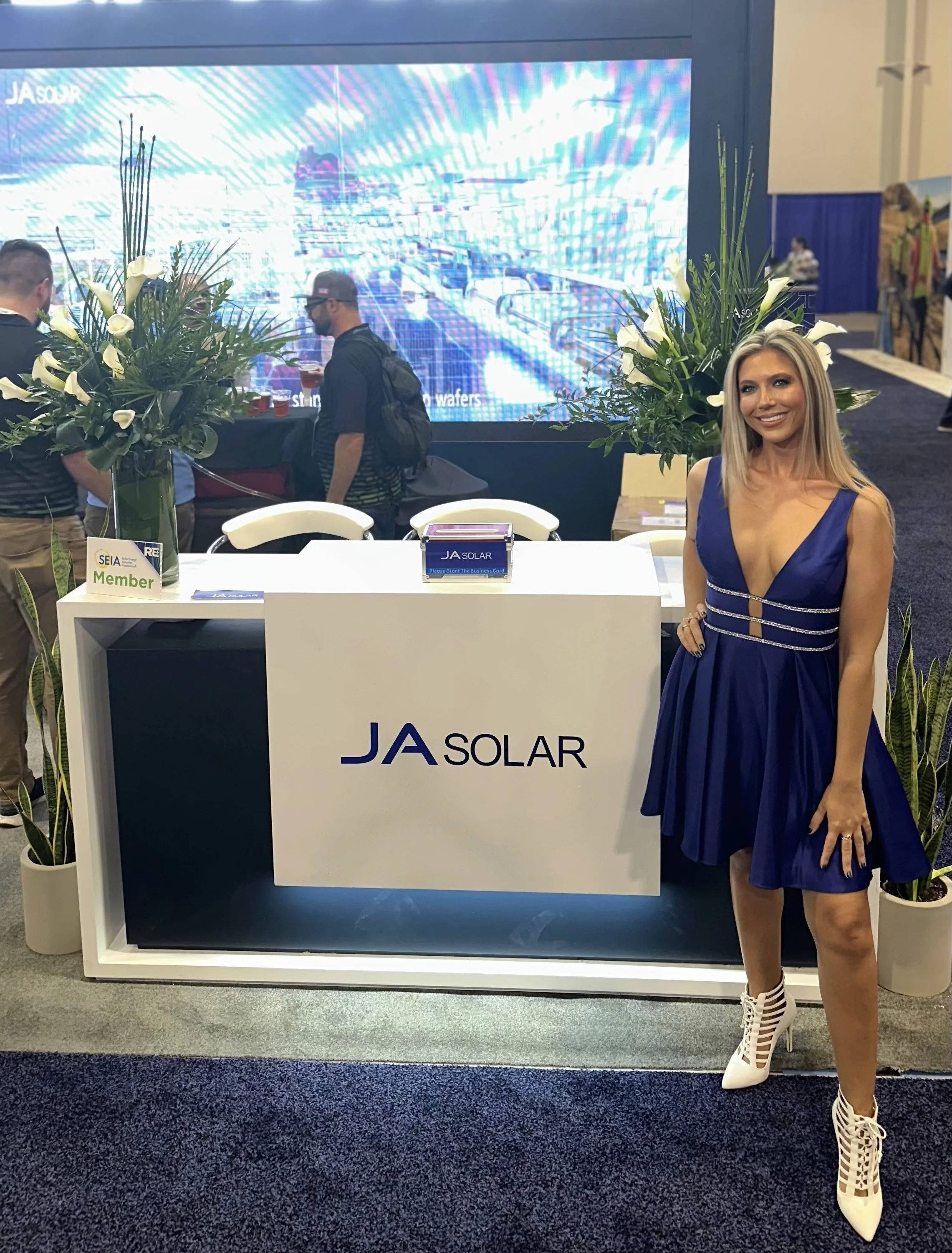 Woman in a blue dress and white high heels standing at a trade show booth for JA Solar, with large floral arrangements and a digital display in the background.