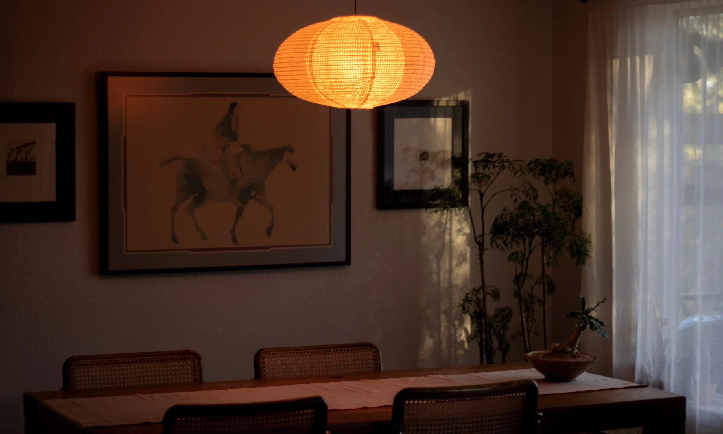 A dining room illuminated by a warm yellow paper lantern hanging from the ceiling, with a wooden table and rattan chairs, framed artwork on the wall, and a plant near a window with sheer curtains.