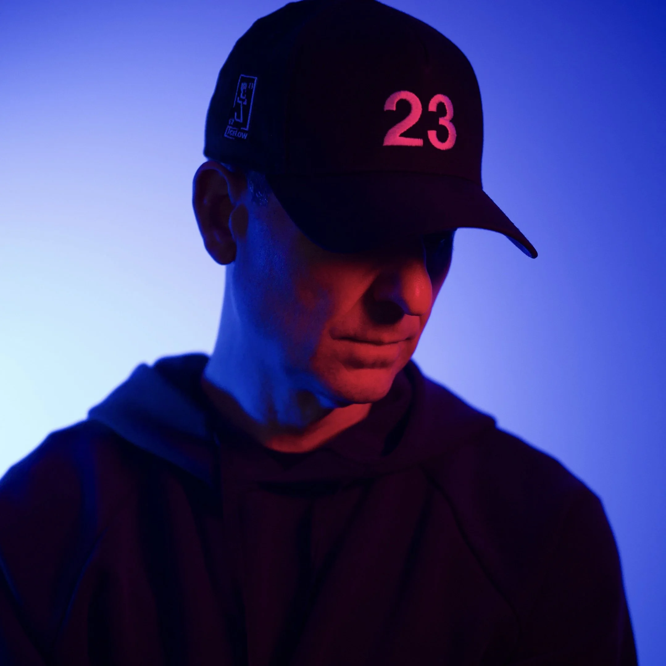 A person wearing a black cap with the number 23 in pink and a blue graphic logo on the side, illuminated by purple and blue neon lights in a dark environment.