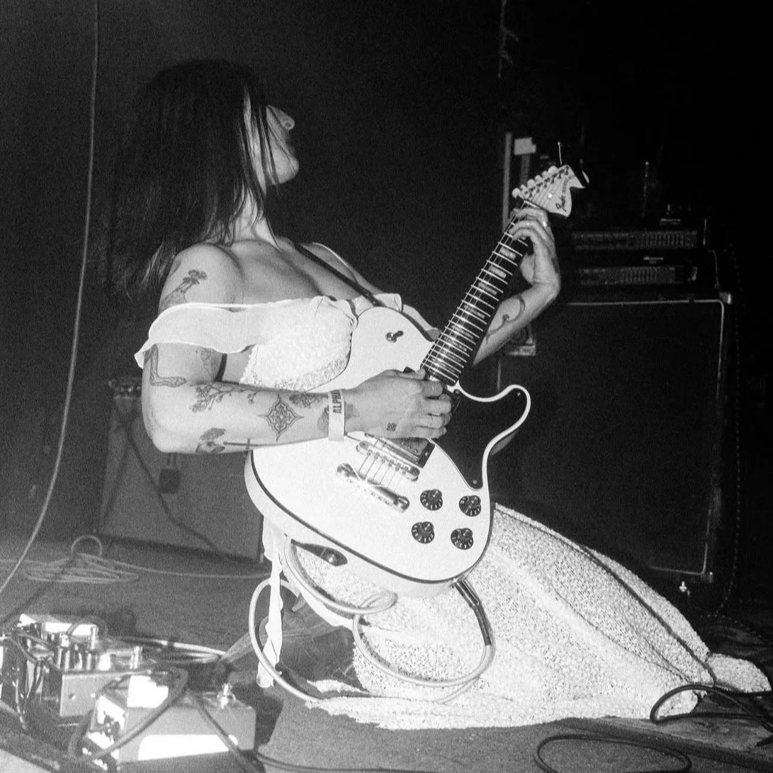 A woman with tattoos playing an electric guitar on stage, with amplifiers and equipment in the background.