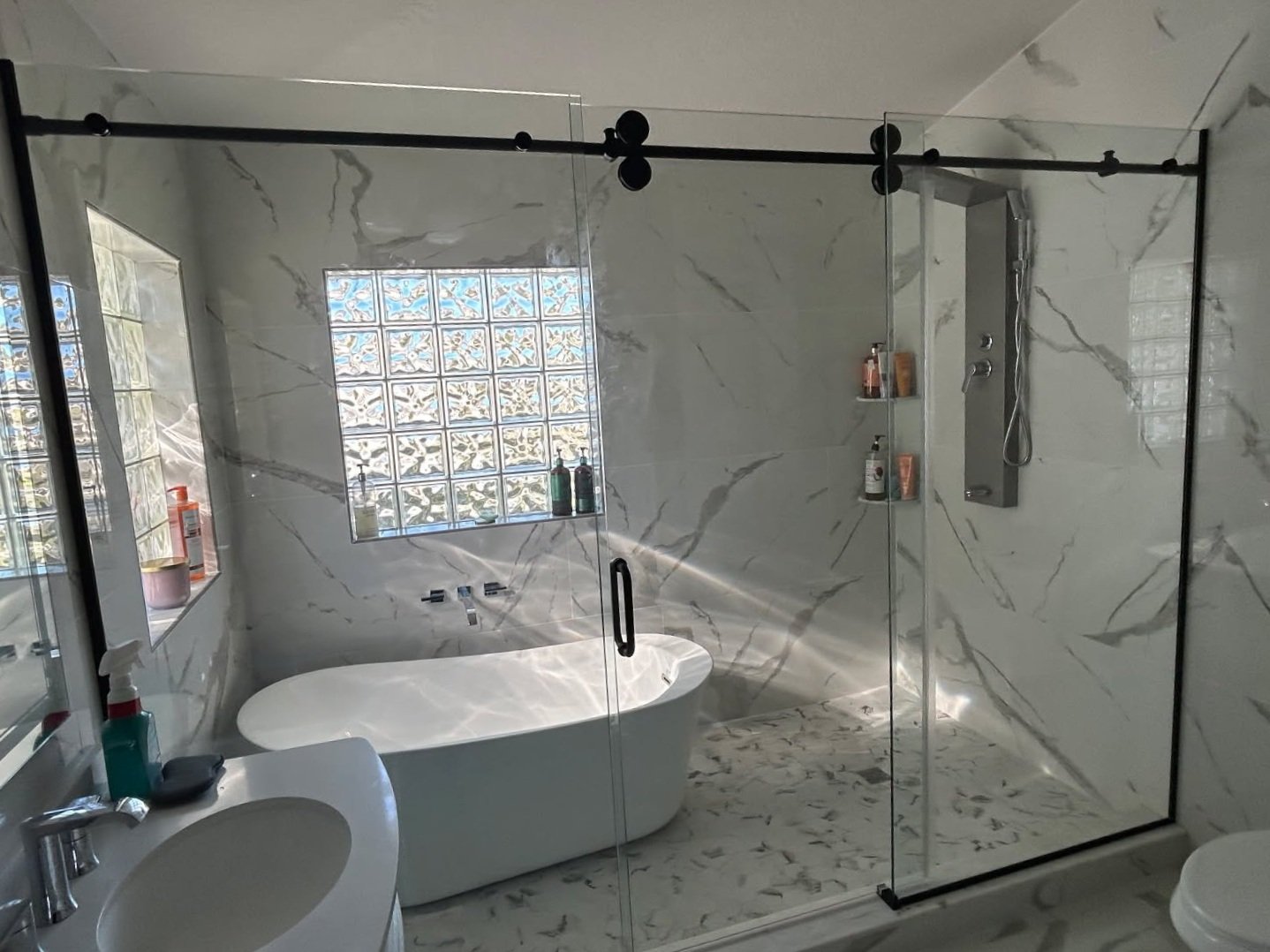 Modern bathroom with marble walls and floor, glass shower enclosure, bathtub, glass block window, and various bath products.