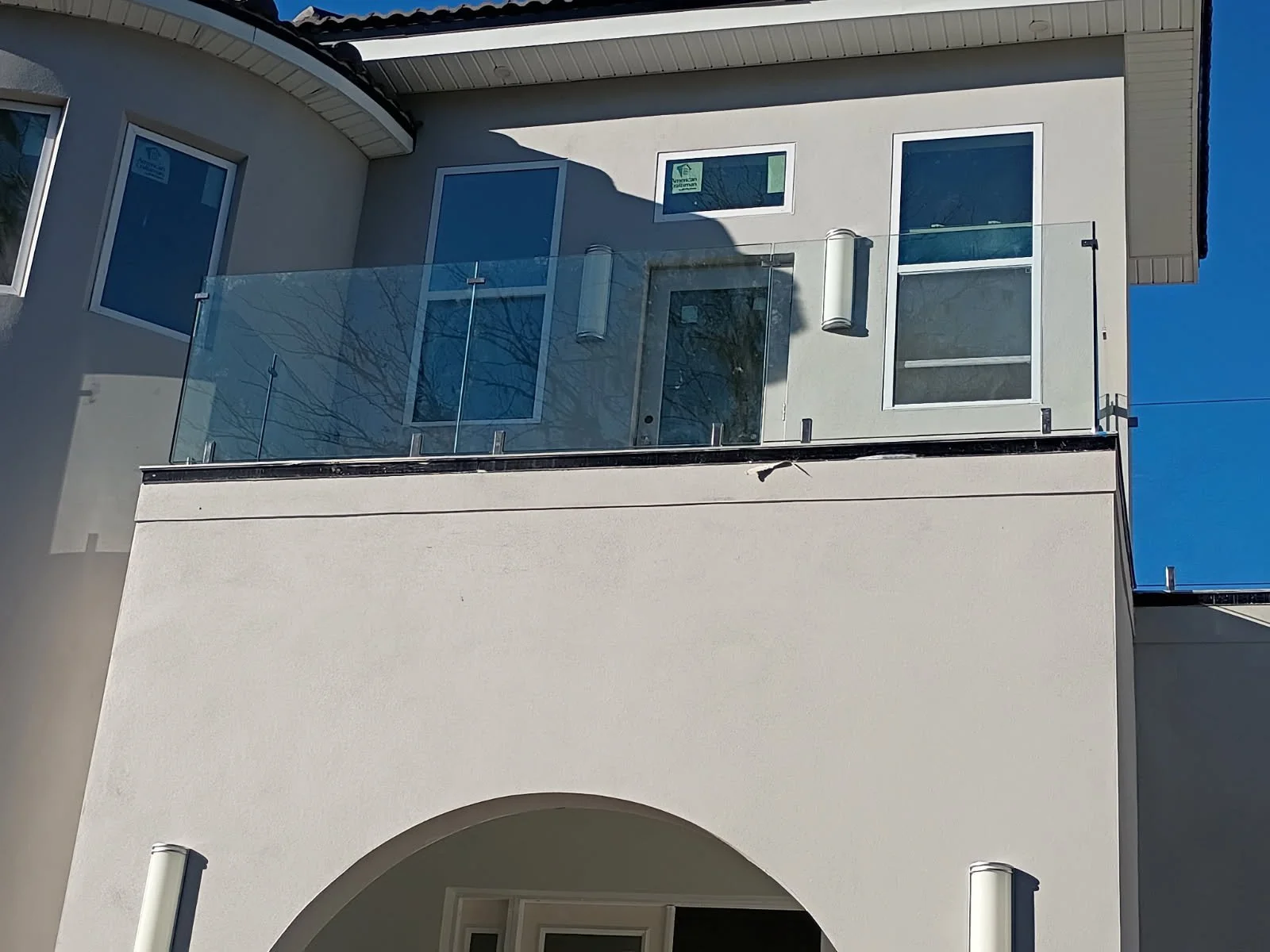 Modern two-story house with large glass windows and balcony, white brick steps, and a green lawn with trees reflected in the windows.