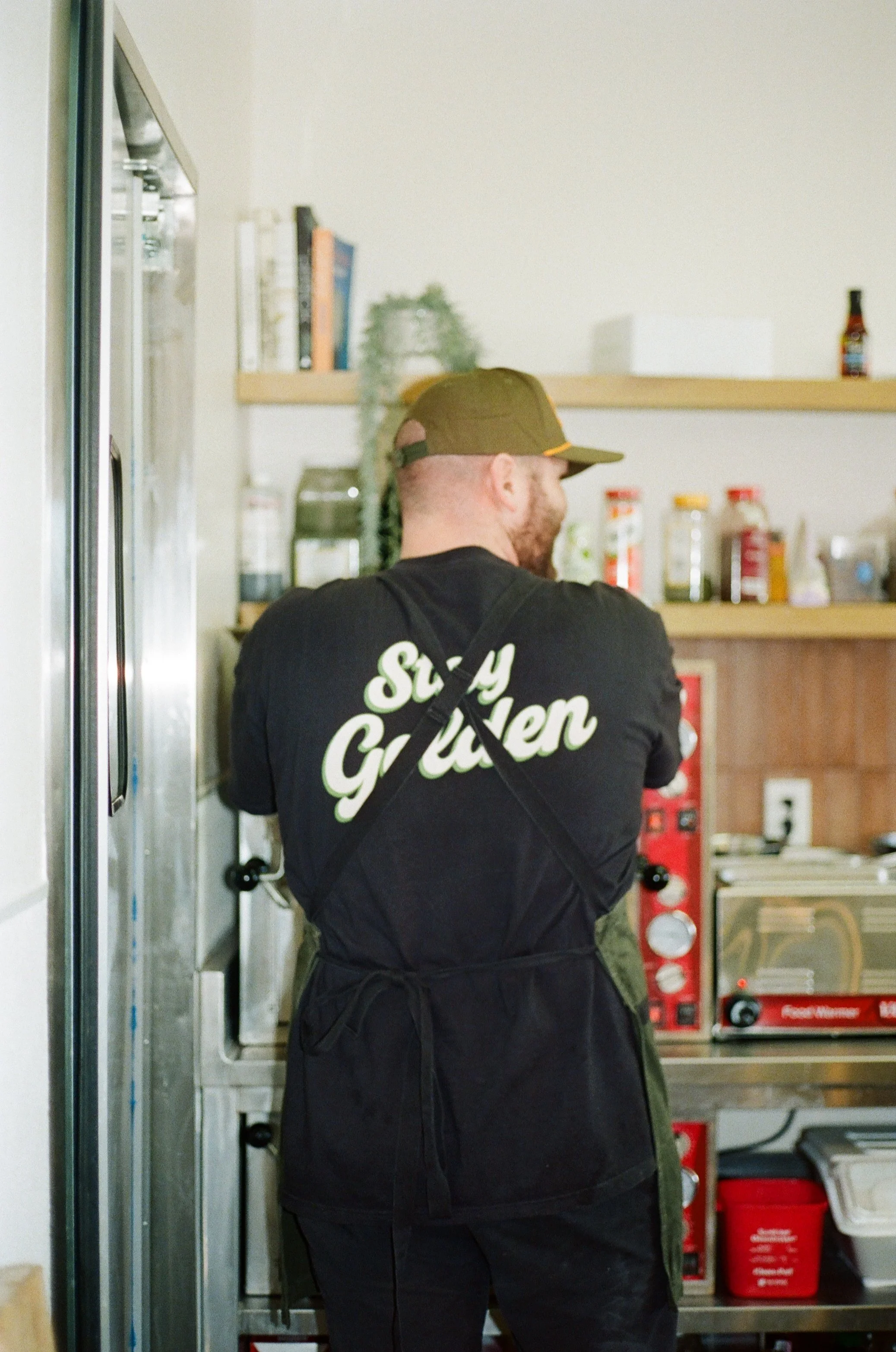 One of the owners of Golden Boy Market & Deli toasting fresh bread in the kitchen, wearing a Golden Boy shirt, hat, and apron while preparing handcrafted sandwiches at the Henderson, NV deli.