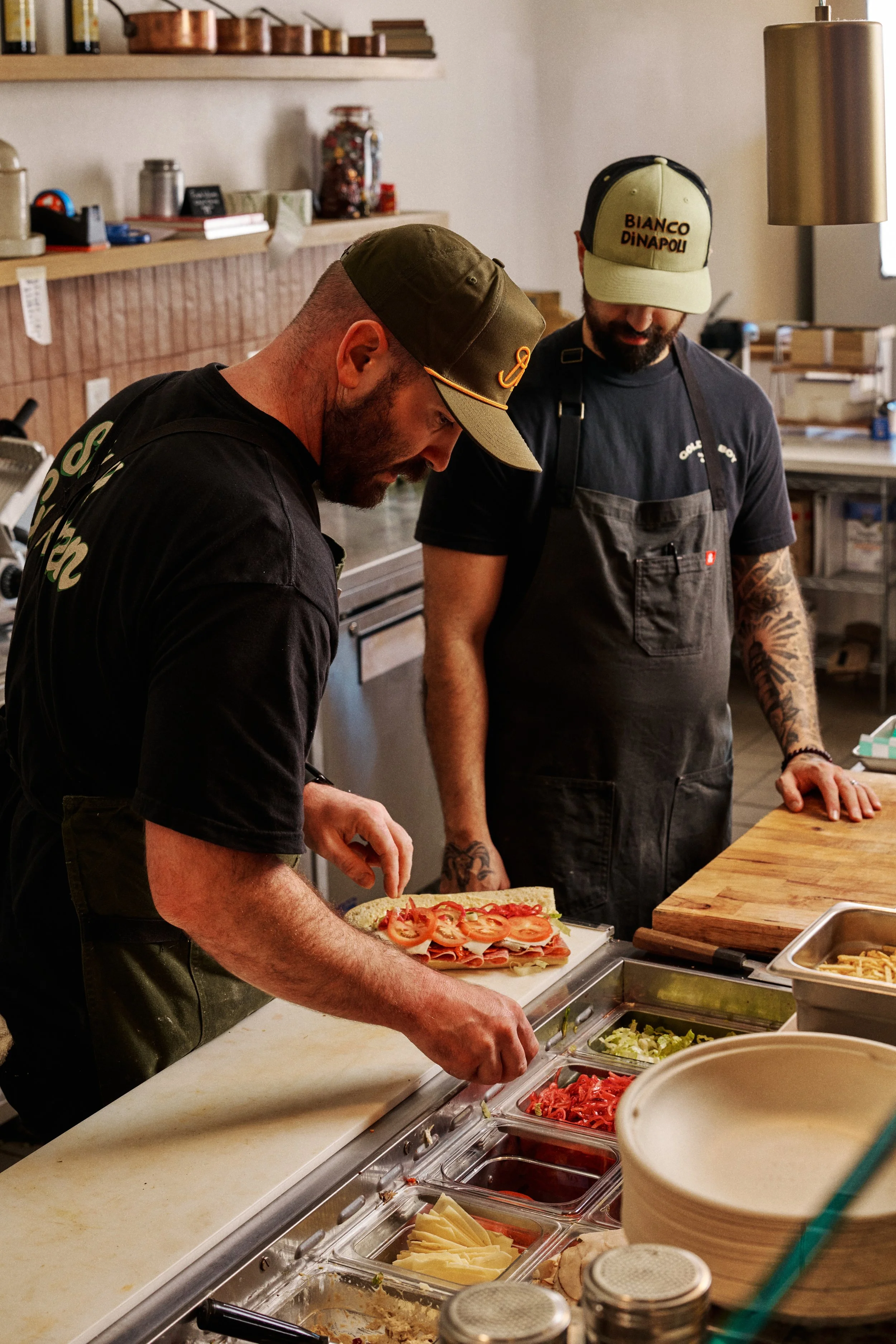 Two men preparing a sandwich with sliced tomatoes, lettuce, and turkey in a deli or sandwich shop.