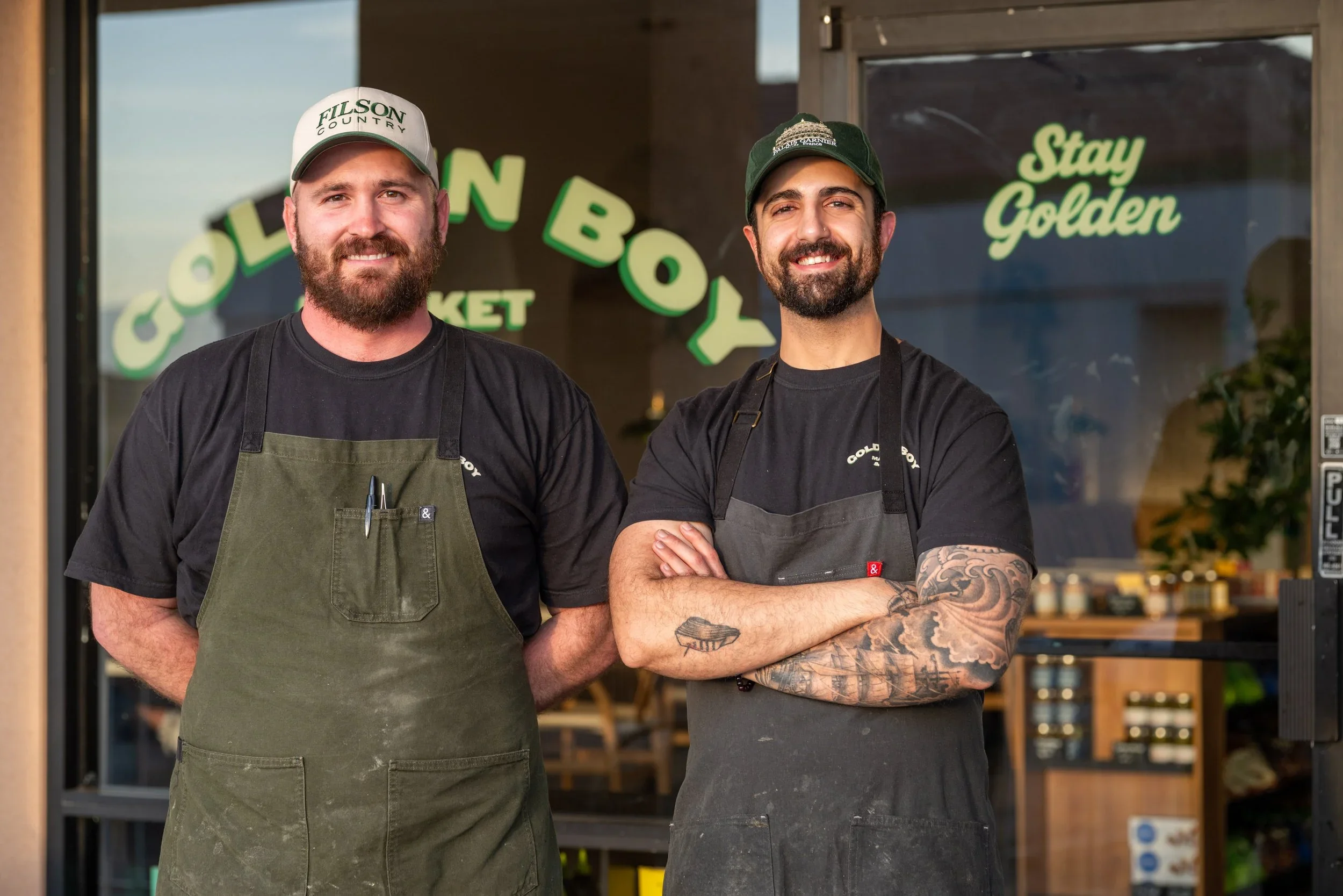 A photo of the owners, the two men are both bearded, wearing hats and aprons. They're smiling outside of Golden Boy deli, behind them is the hand painted sign