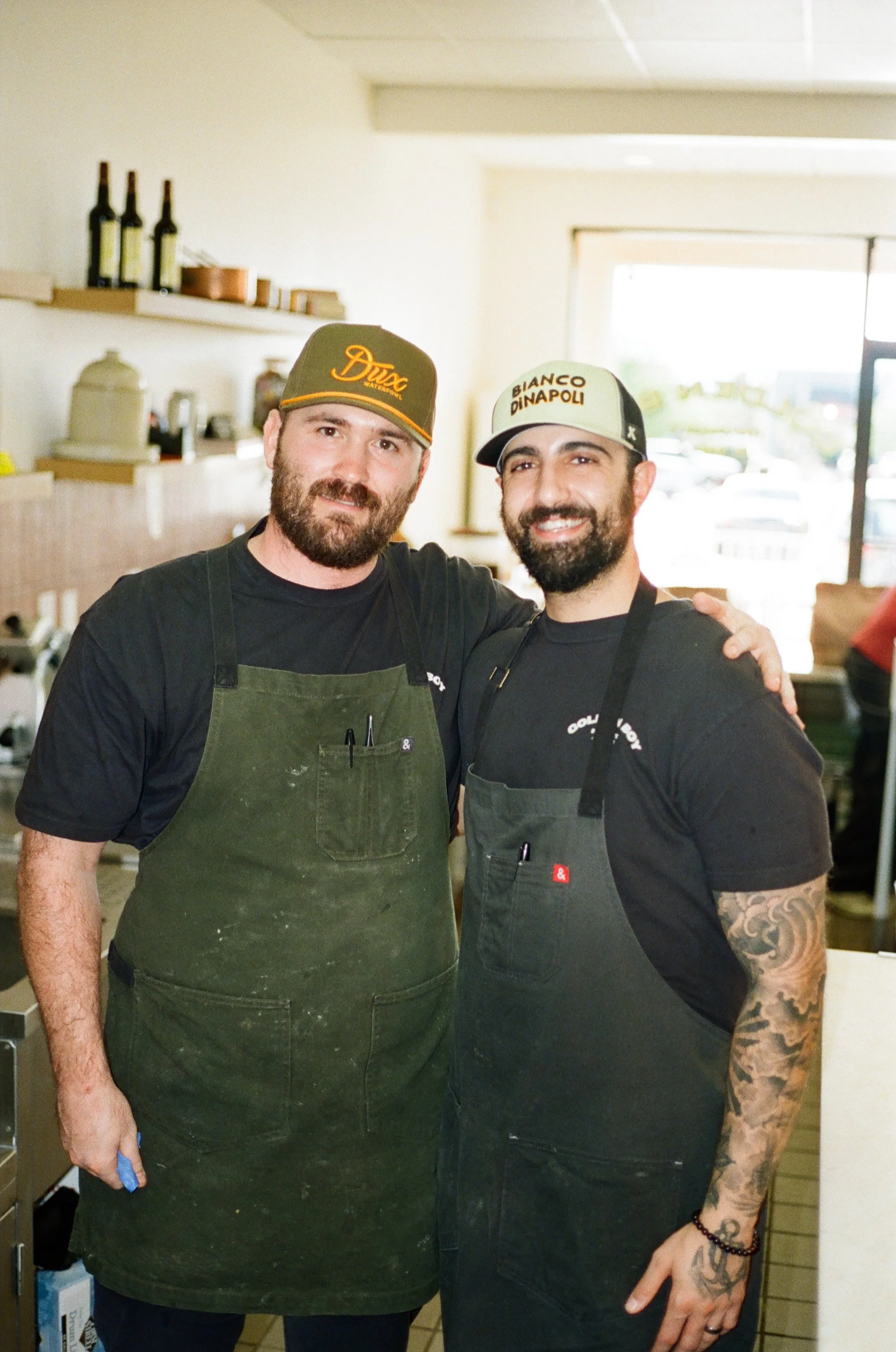 Two men wearing aprons and hats, smiling inside a restaurant or cafe. One has a beard and is wearing a baseball cap, the other has tattoos on his arm and is also wearing a baseball cap.
