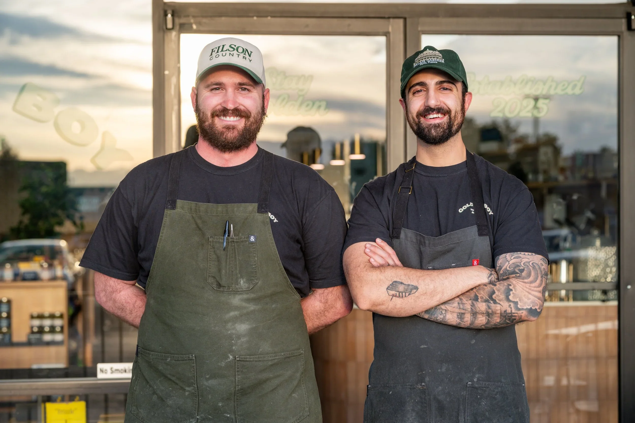 Two smiling men standing in front of a glass door, wearing black t-shirts and aprons, with one wearing a gray baseball cap and the other a green cap. They are outside Golden Boy deli, with a reflection of their handprinted sign.