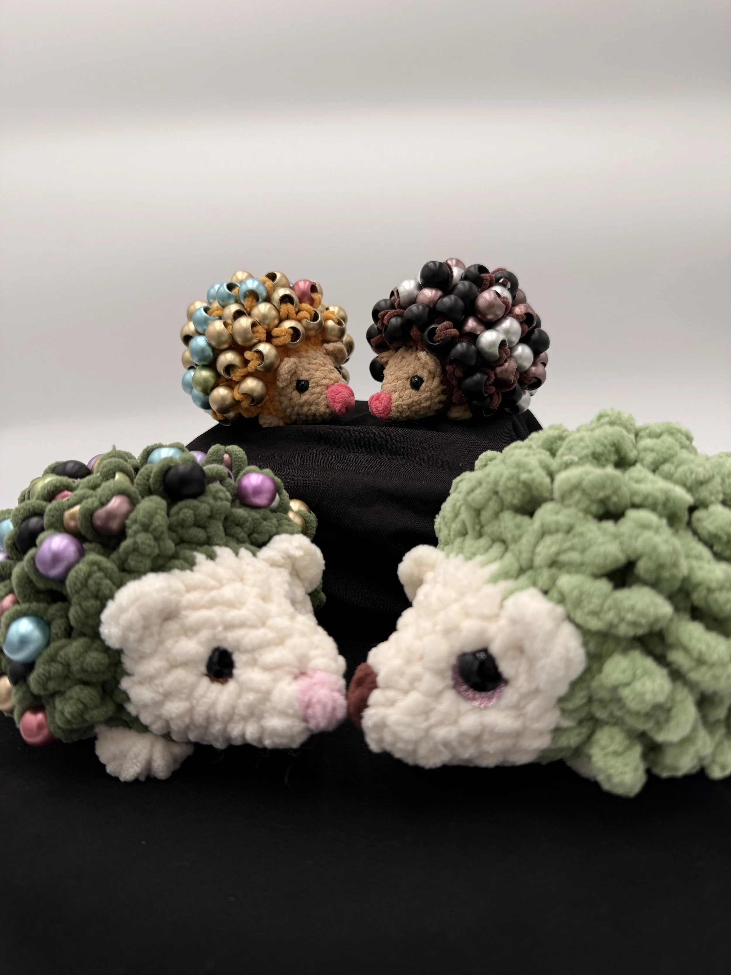 Two handmade plush animals with curly, bead-filled hair, sitting on black fabric, surrounded by more plush animals, some with bead embellishments.