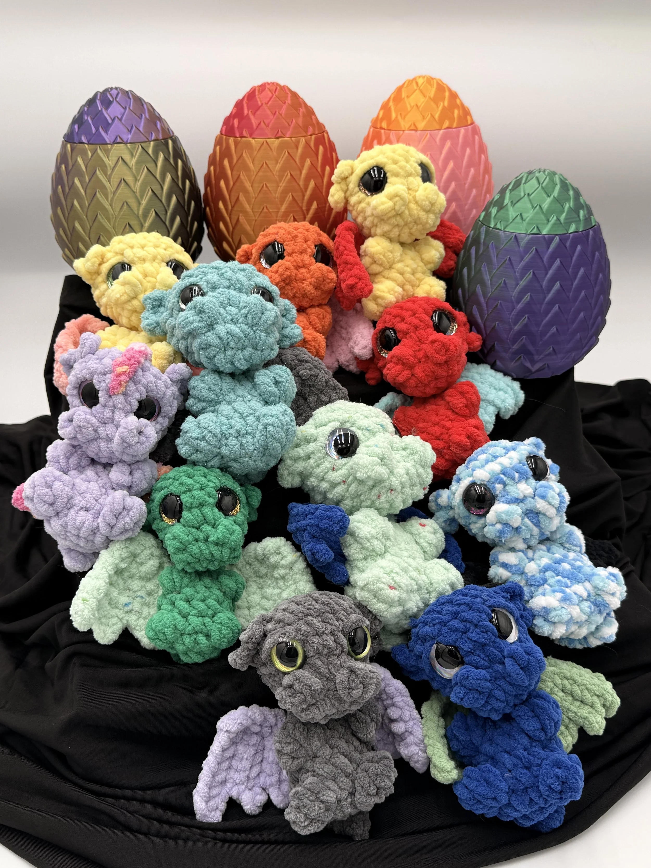 Colorful plush animal toys resembling clouds with large black eyes, arranged with decorative eggs in the background. The toys are in various colors like purple, green, blue, gray, red, orange, yellow, pink, and multicolored, displayed on a black fabr