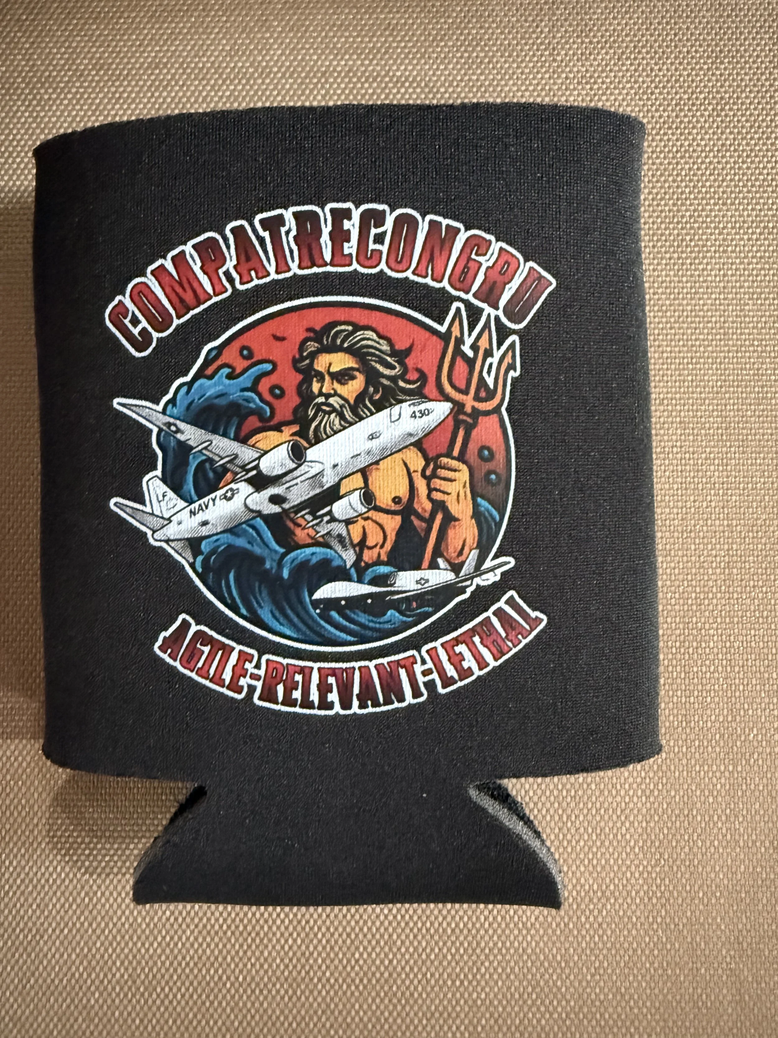 A black fabric wristband with a colorful military-style patch featuring a muscular bearded man holding a jet fighter, surrounded by water and flames, with the text 'COMPATRECONGRU' at the top and some additional text at the bottom.