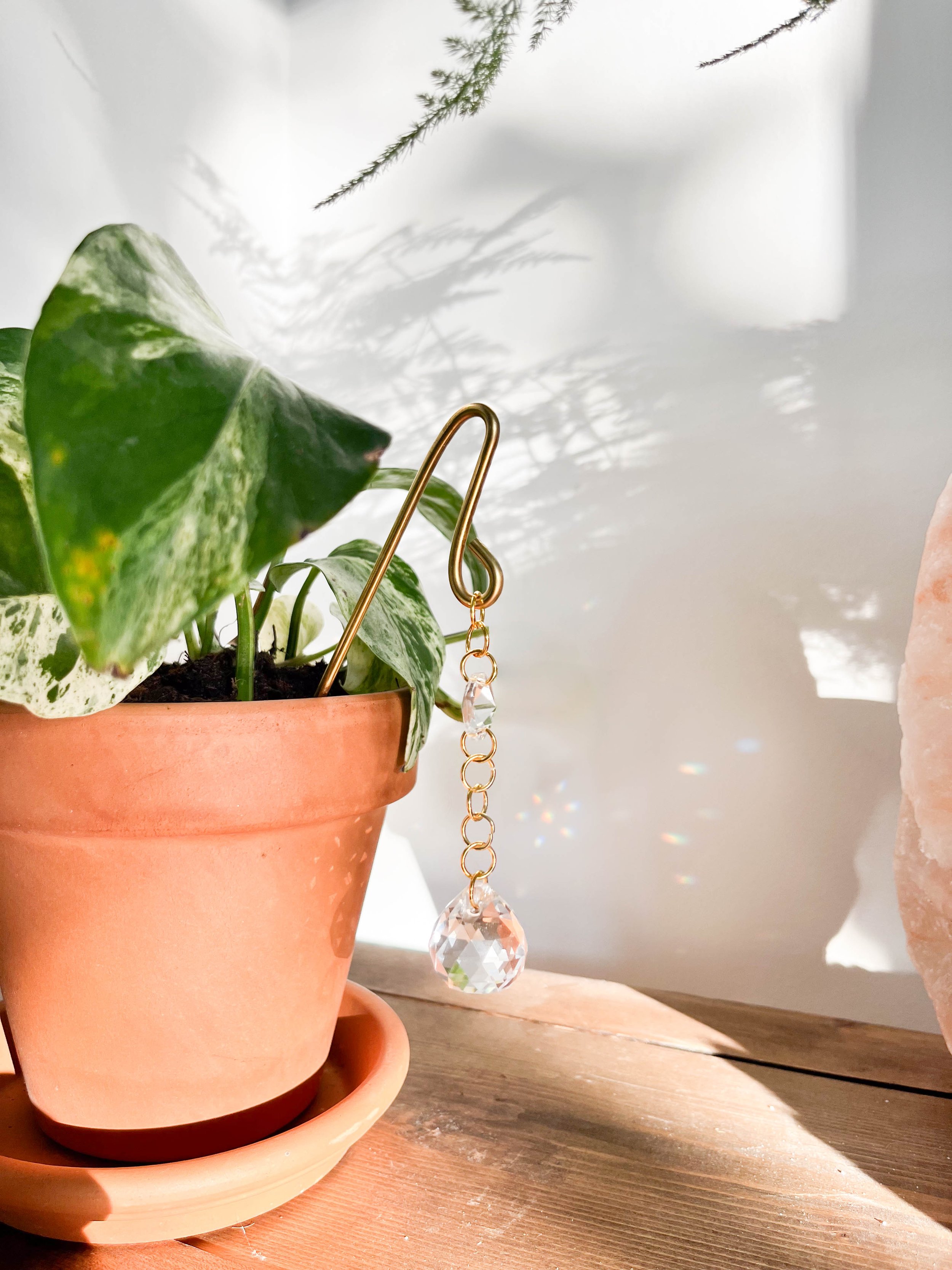 Gold plant pot charm with a delicate gold chain featuring a small glass prism above and a large glass prism hanging below, designed to reflect sunlight and brighten your indoor garden.
