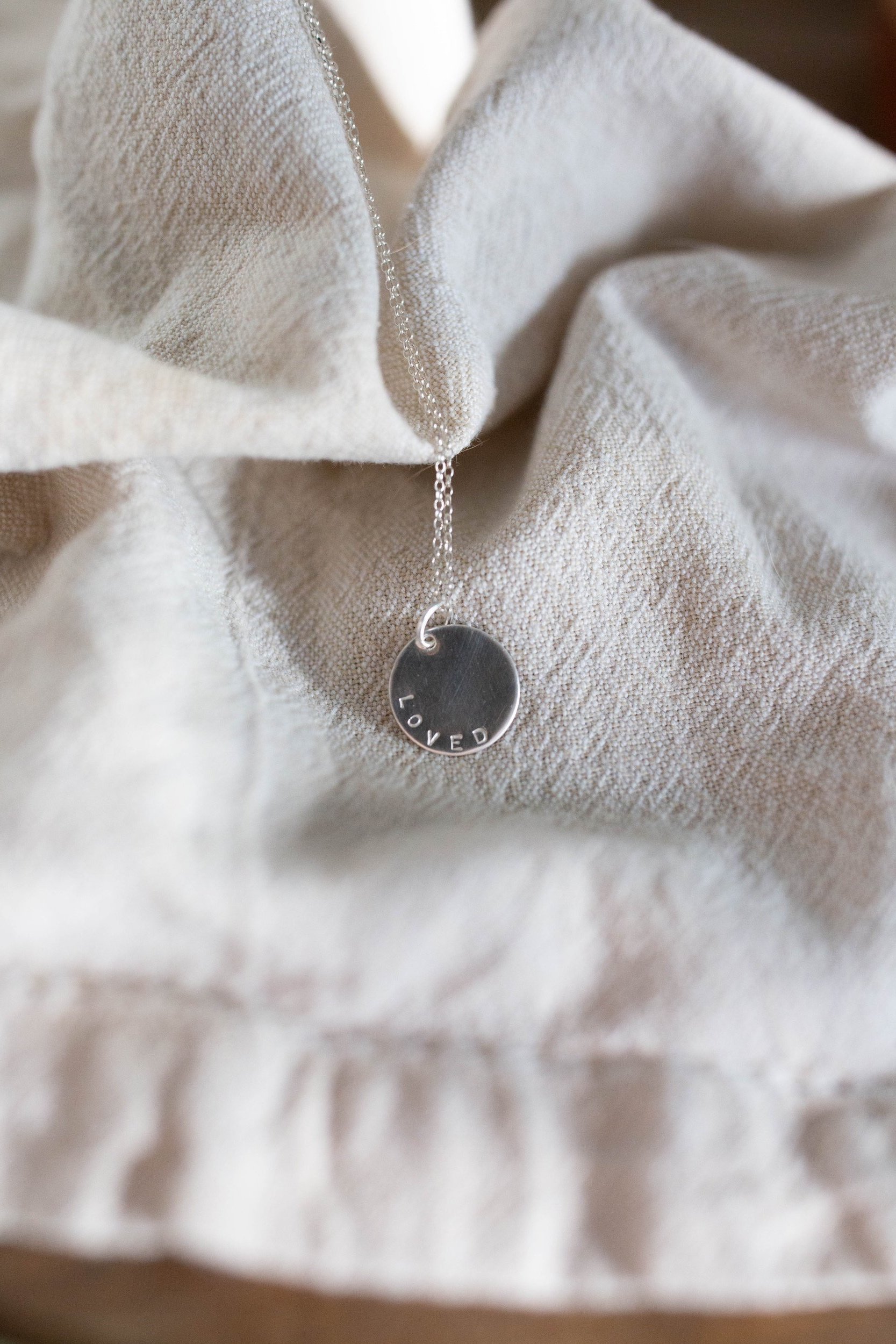 Angled view of sleek silver affirmation necklace featuring 'Loved' mantra. Handcrafted pendant catches soft light, emphasizing the stamped detail. Modern, intentional jewelry design