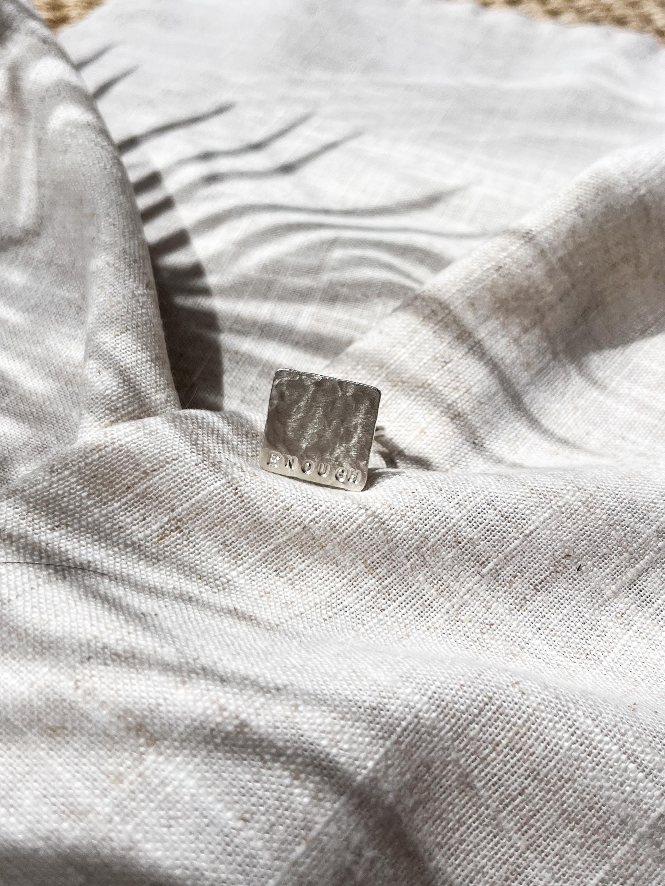 Top view of silver square textured ring hand stamped with the word "Enough," displayed on linen fabric in warm sunlight. Minimalist affirmation jewellery with a meaningful message.