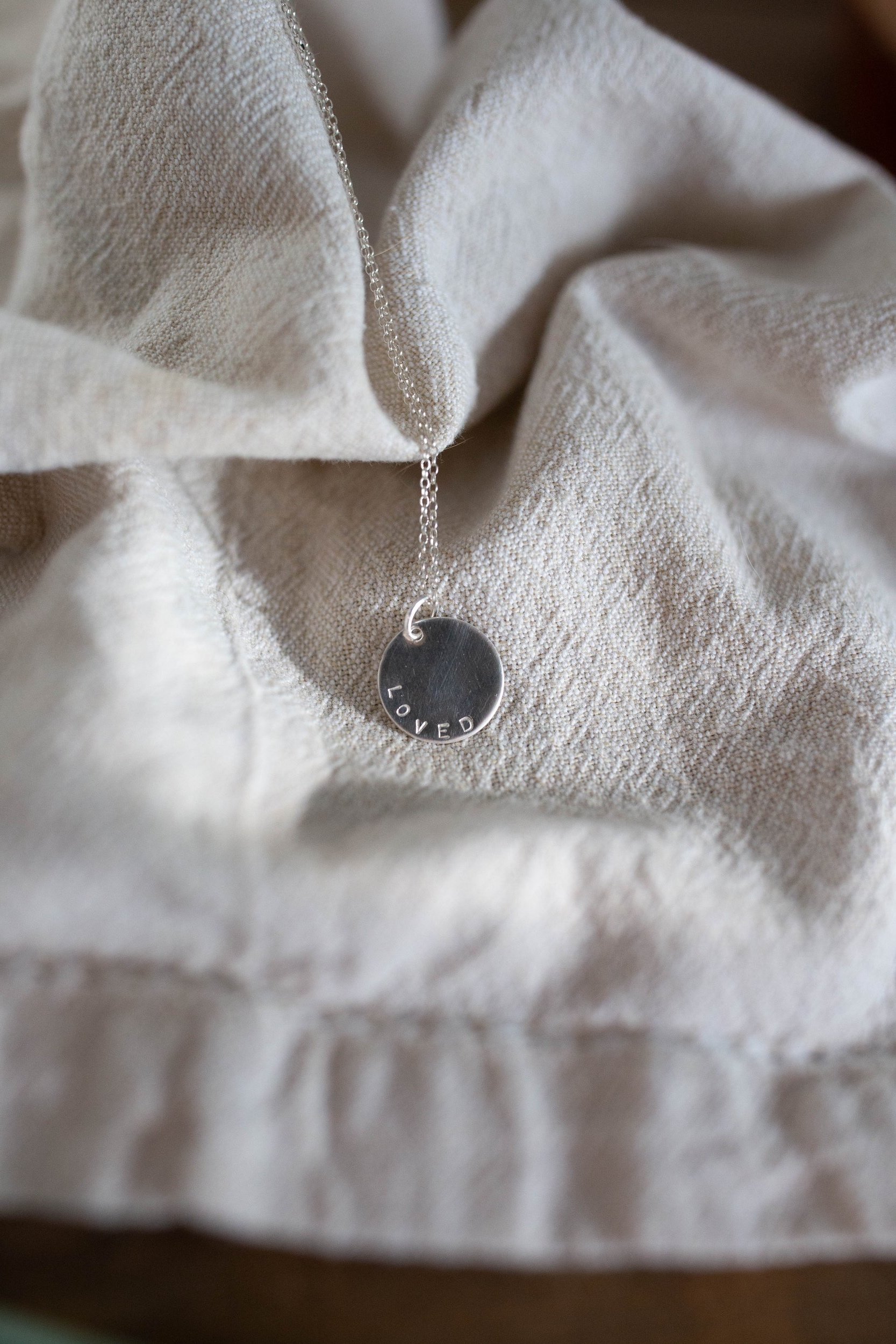 Emotive lifestyle shot of 'Loved' silver charm necklace on fabric with soft shadows. Thoughtful self-love or encouragement gift, handmade from eco-conscious sterling silver.