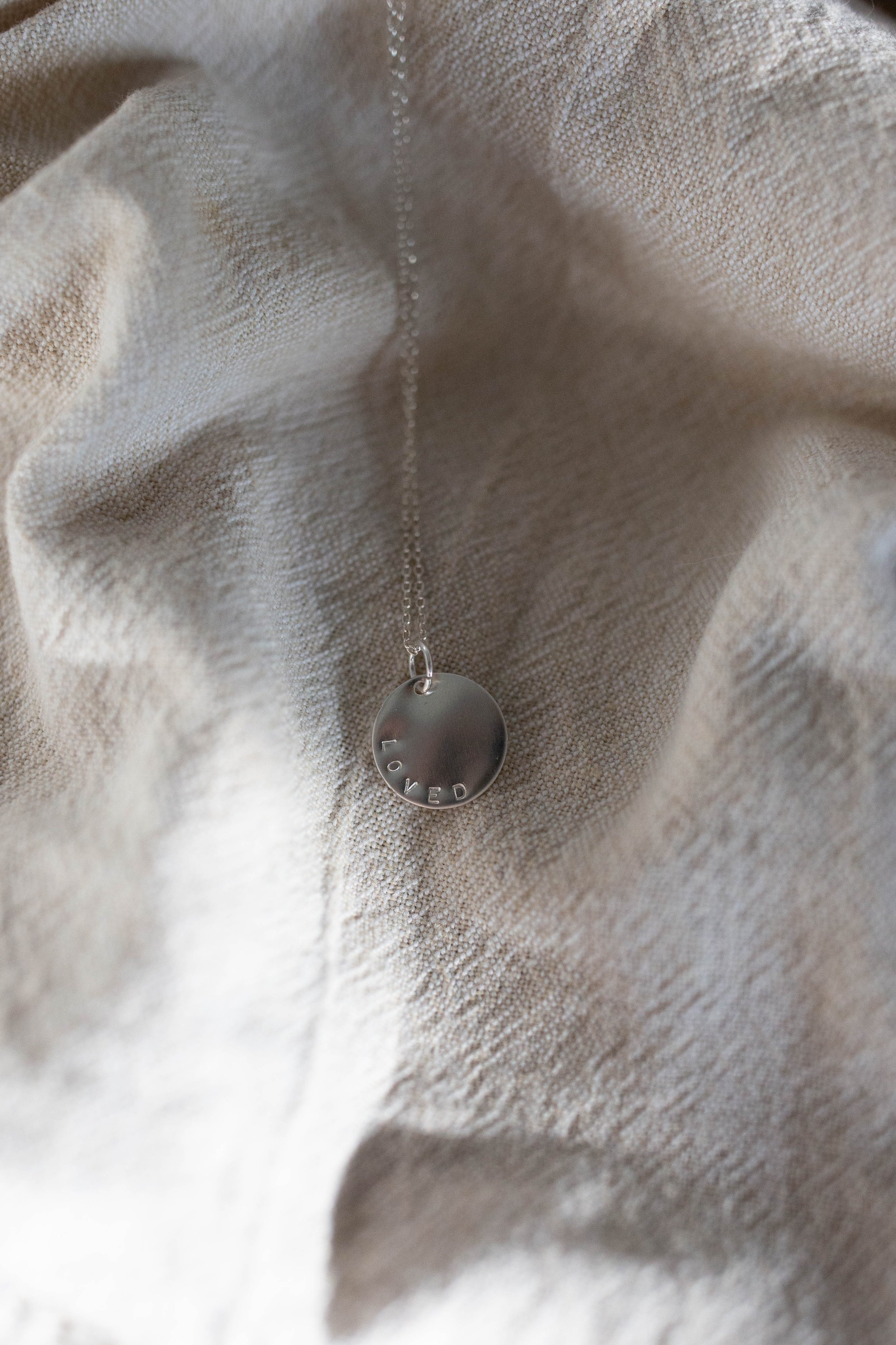 Close-up of personalized silver disc pendant stamped with the word 'Loved.' Elegant handcrafted jewelry photographed in dappled sunlight on linen. Ideal for custom affirmation gifts