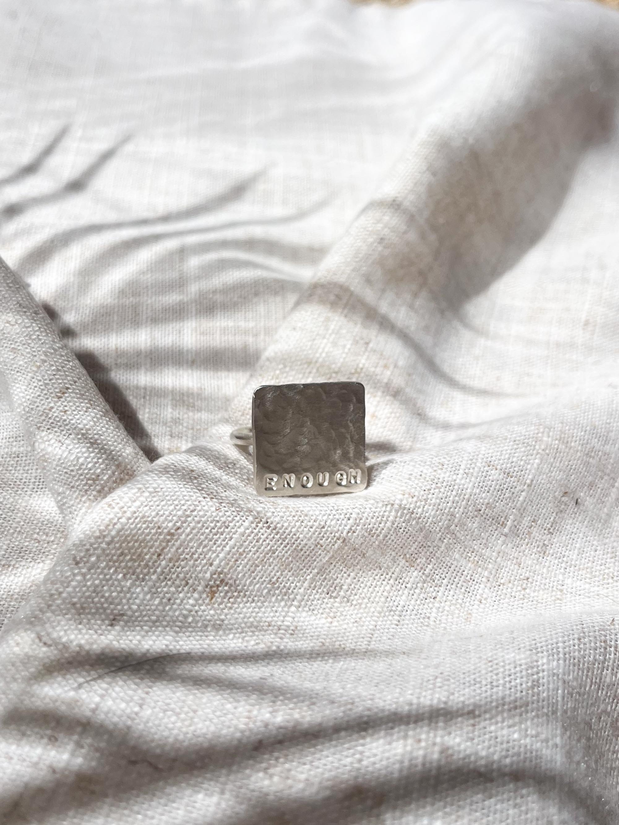 Angled shot of silver affirmation ring with "Enough" hand stamped on its edge. Resting on linen in golden sunlight, this image shows off the modern square design and artisanal texture.
