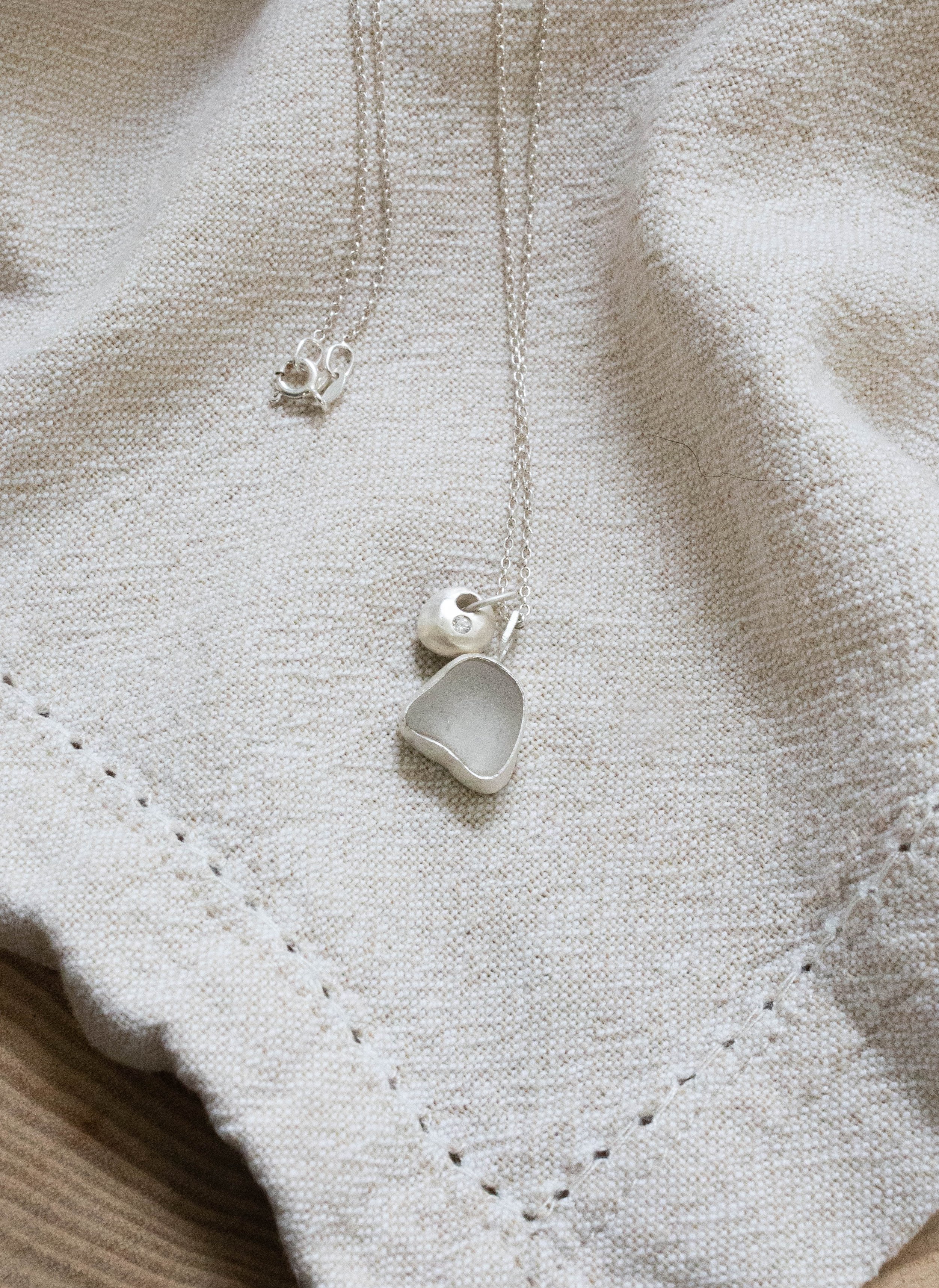 Close-up of clear Cornish sea glass and silver pebble charm with CZ stone on linen. Ocean-inspired handmade necklace crafted with eco-friendly recycled silver.