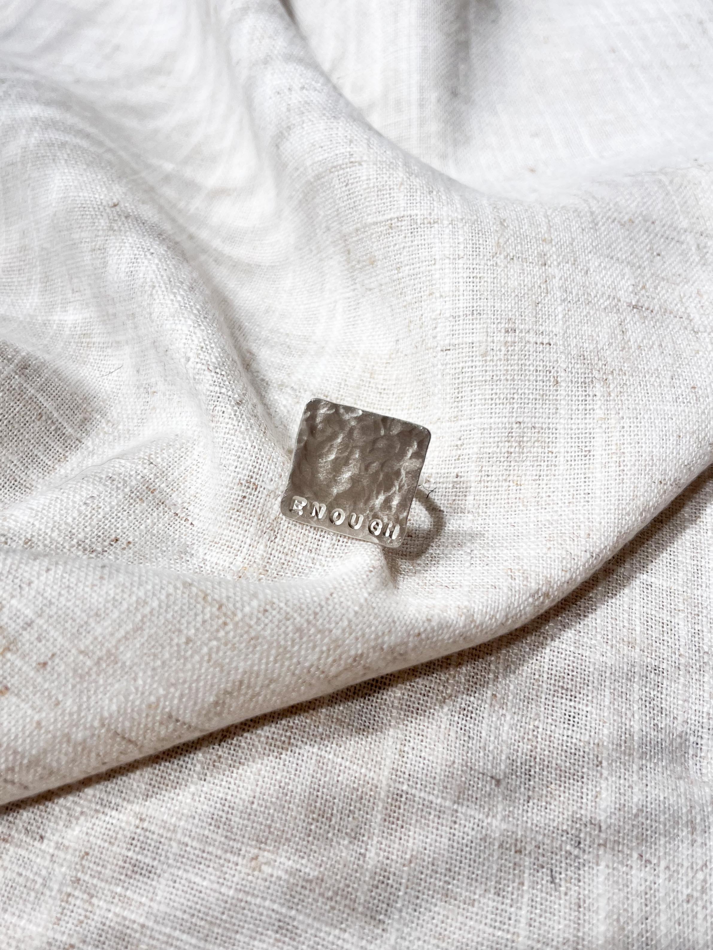 Close-up of the word "Enough" stamped along the edge of a handcrafted silver textured ring. Captured on a soft linen backdrop with natural sunlight, highlighting the detailed finish.
