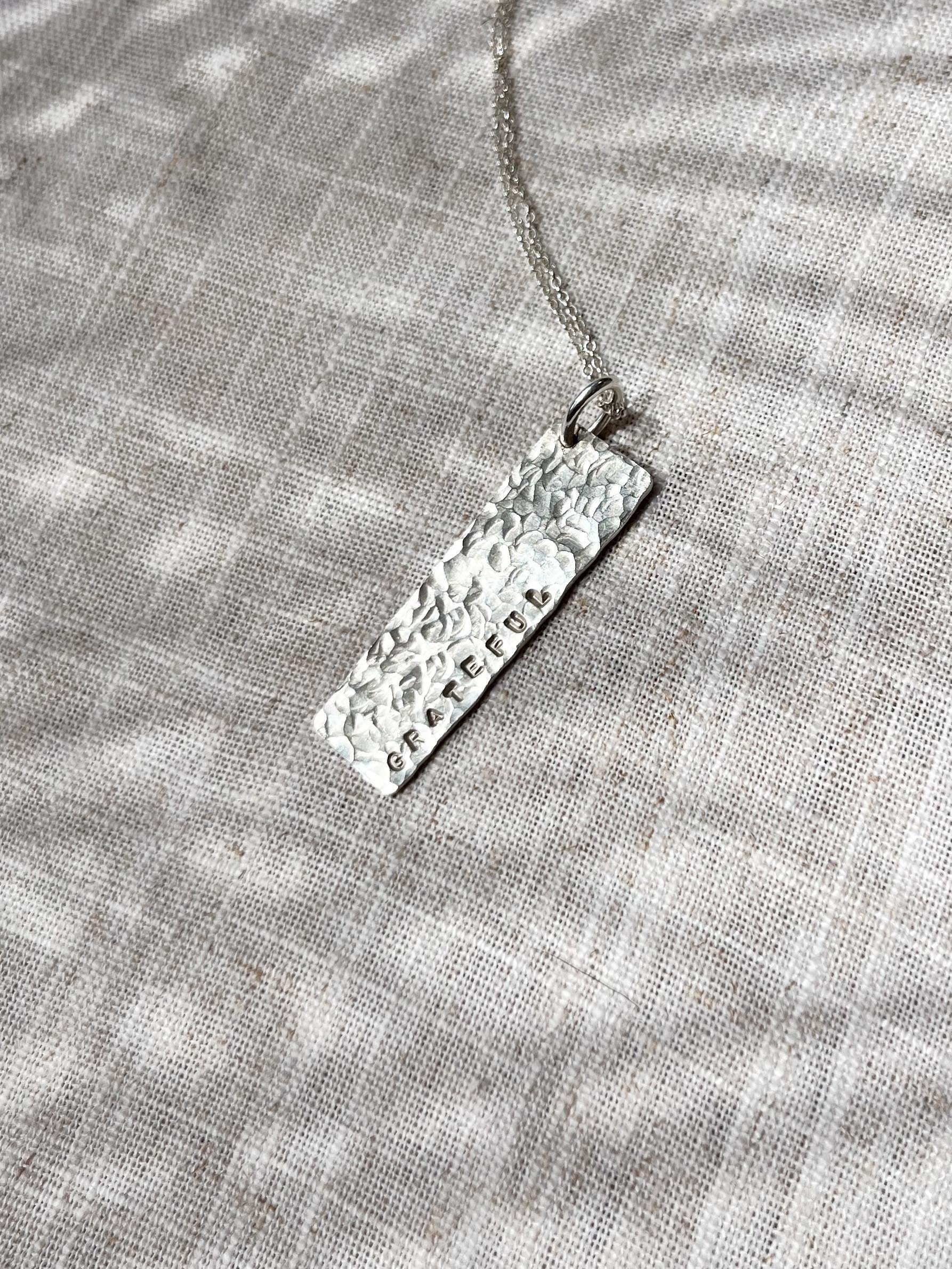 Close-up of textured rectangular silver tag necklace hand stamped with the word "Grateful," photographed on fabric in natural sunlight. Minimalist personalised jewellery with meaningful affirmation