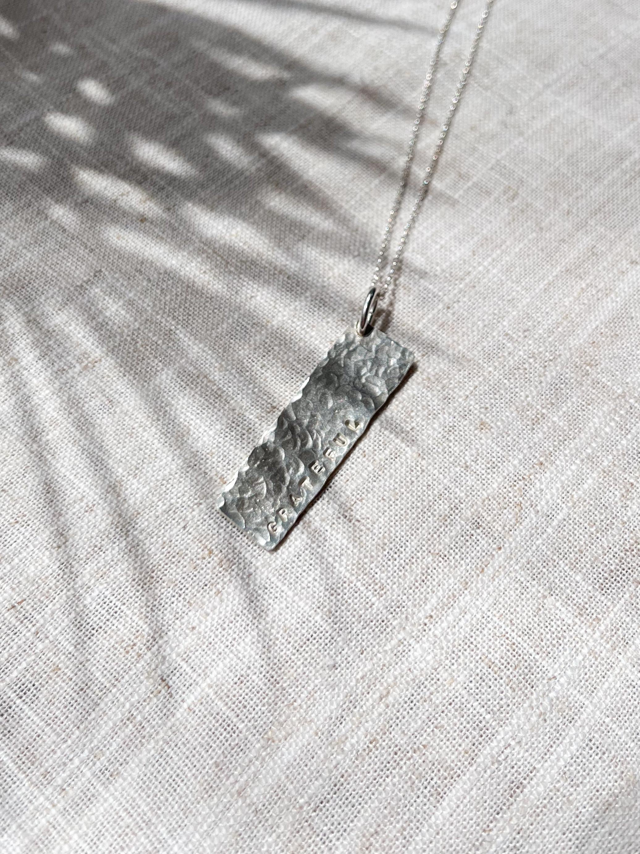 Detailed shot of sterling silver affirmation bar pendant featuring "Grateful" hand stamped on textured tag, resting on fabric with sunlight highlighting craftsmanship and texture.