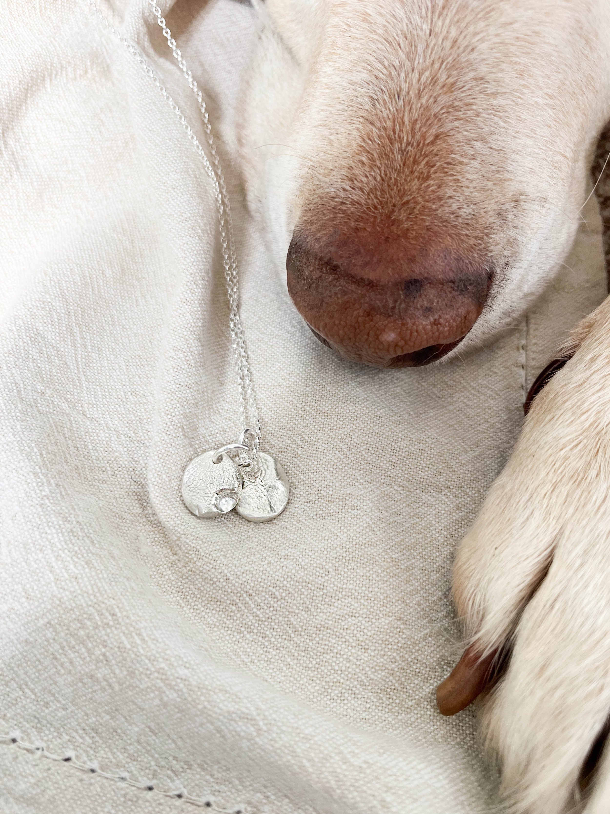 Handmade silver dog nose print pendant resting on linen with the dog’s nose and paw visible. A heartfelt custom pet keepsake and memorial gift, created using your dog’s actual nose print.
