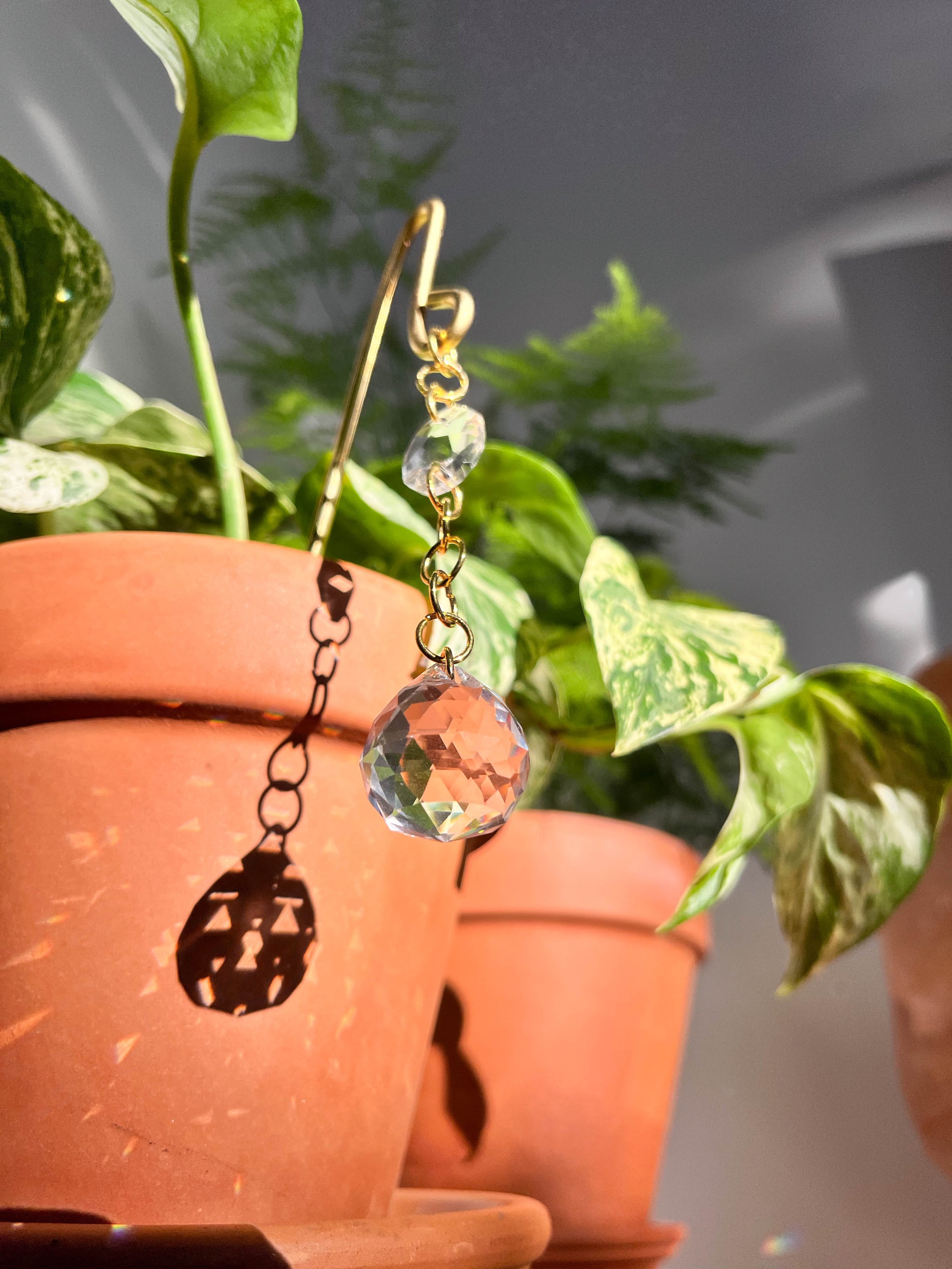 Handmade plant pot charm with gold chain and dual glass prisms hanging delicately, creating vibrant light reflections in indoor gardens.