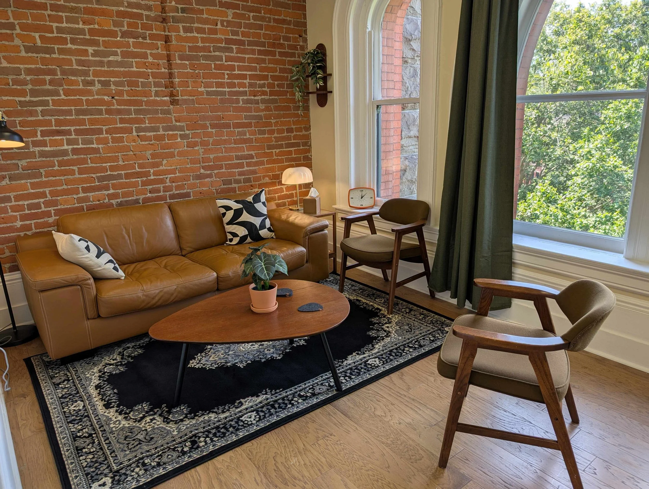 room with a tan leather sofa, patterned pillows, a dark rug, a wooden coffee table with a potted plant, two wooden and upholstered chairs, a side table with a retro clock and lamp, a brick wall, large window with green curtains, and hardwood flooring.