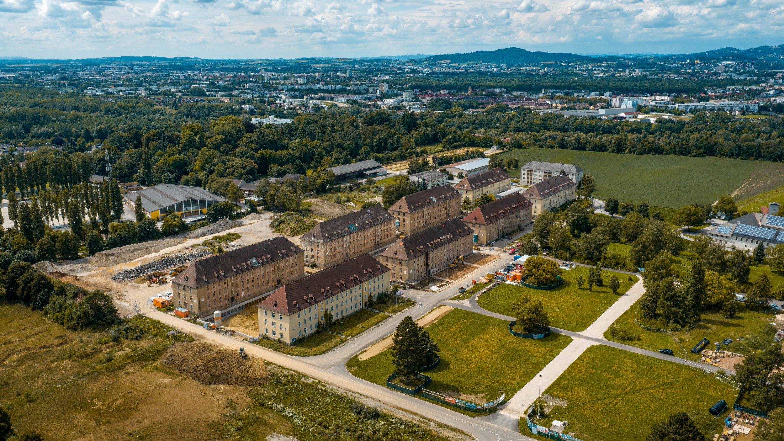 Luftaufnahme eines Wohnkomplexes im Bau mit mehreren mehrstöckigen Gebäuden, umgeben von grünen Feldern, Straßen und Bäumen, mit einer Stadt im Hintergrund.