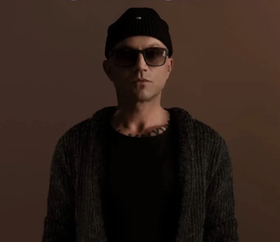 Ein Mann trägt eine schwarze Mütze, dunkle Sonnenbrille, einen schwarzen Pullover und eine dunkle Jacke, vor einem braunen Hintergrund.