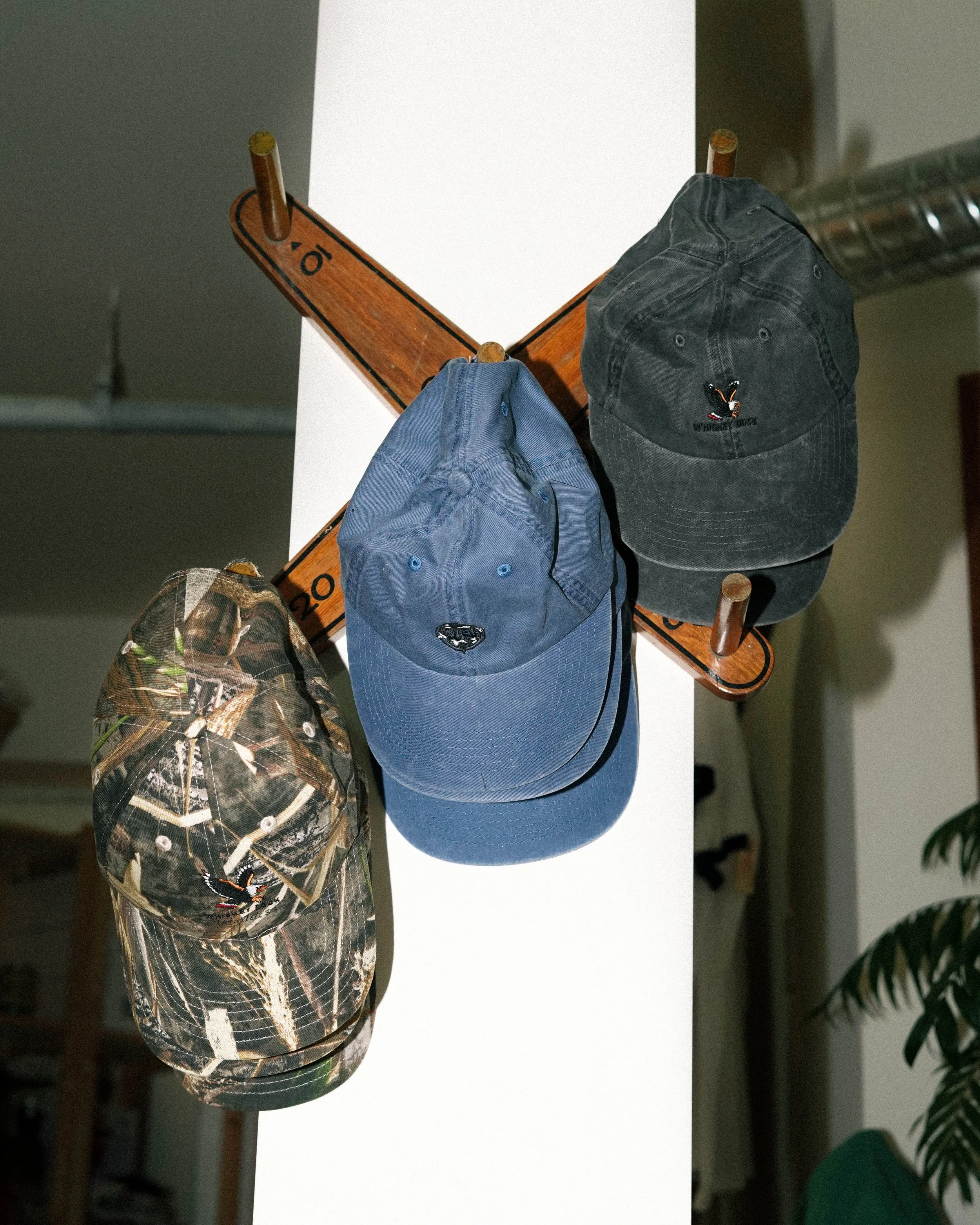 Three baseball caps hanging on a wooden coat rack mounted on a white wall, one with camouflage pattern, one blue, and one black with a small logo.