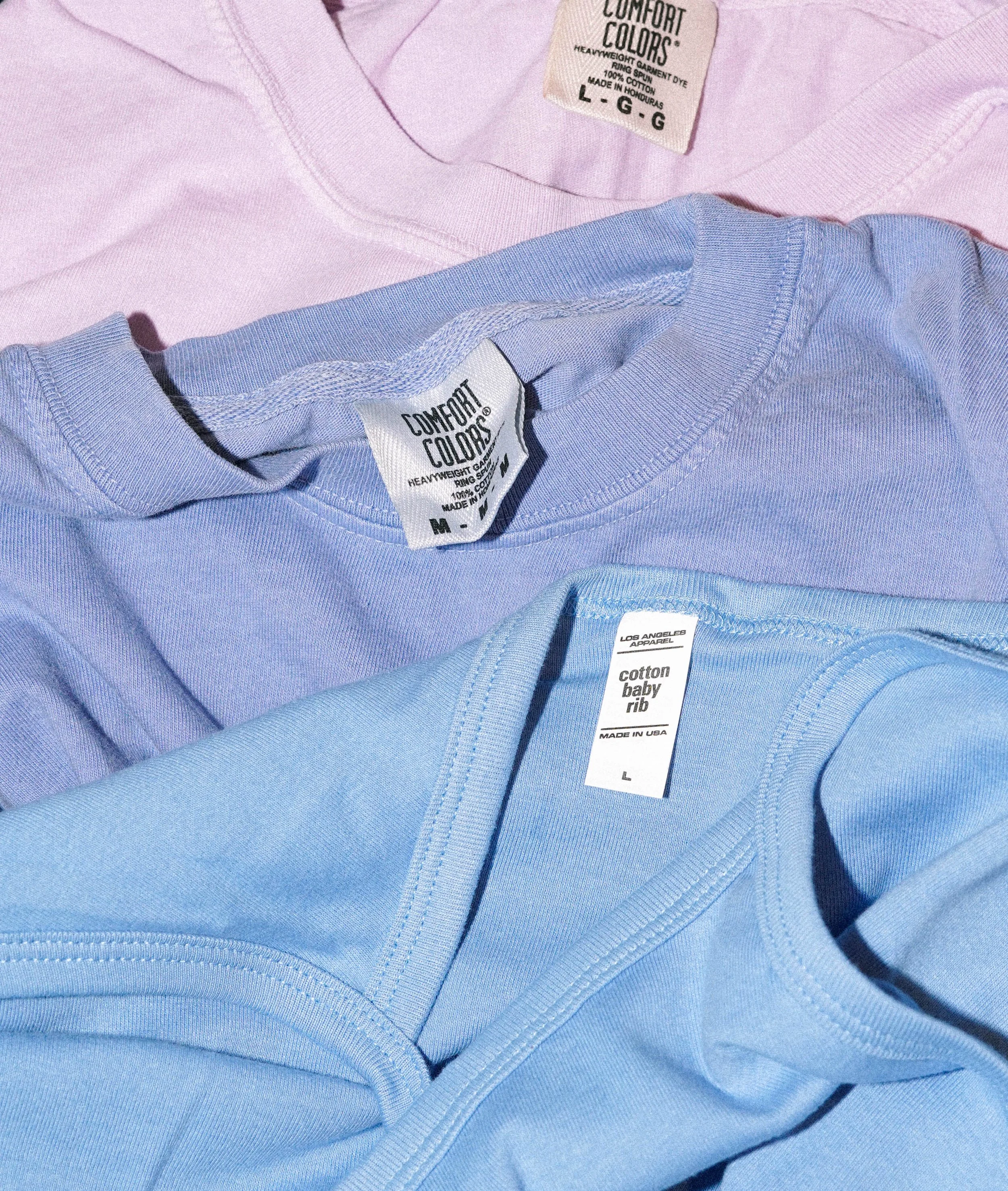 Three folded T-shirts in pastel pink, lavender, and light blue colors with visible tags.