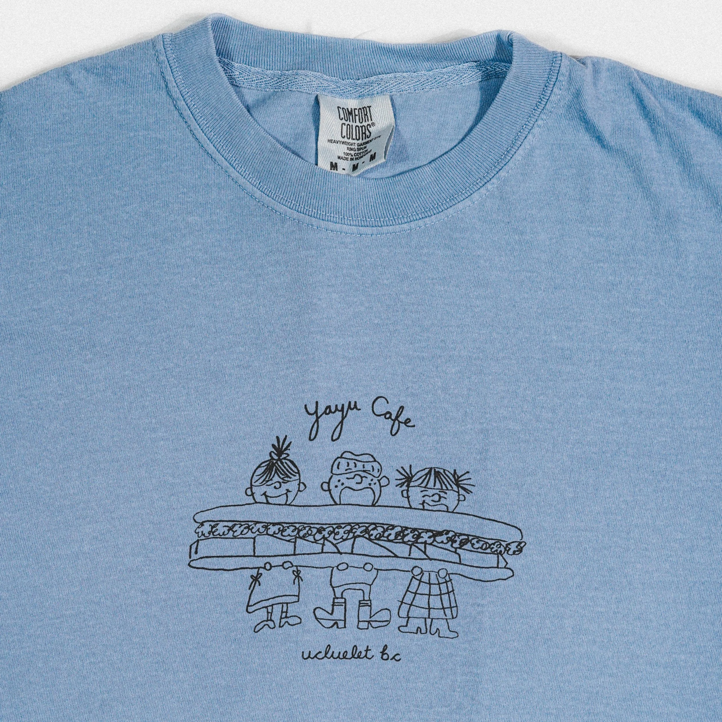 Light blue T-shirt with a black line drawing of three characters sitting at a table labeled "Yayu Cafe" and "Ucluelet BC" underneath.