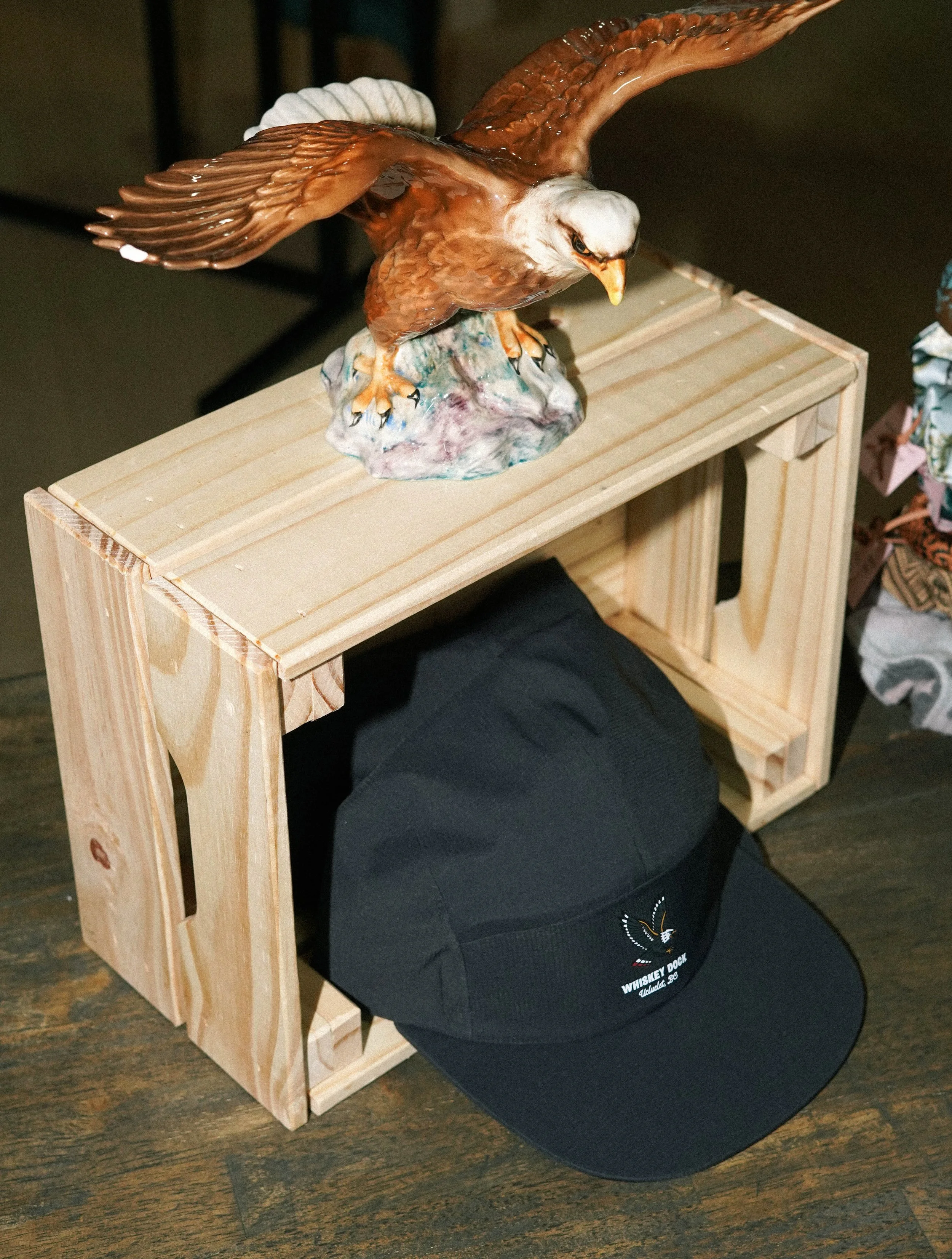 A decorative ceramic eagle sculpture on a wooden crate, with a black baseball cap underneath. The crate is placed on a wooden floor.