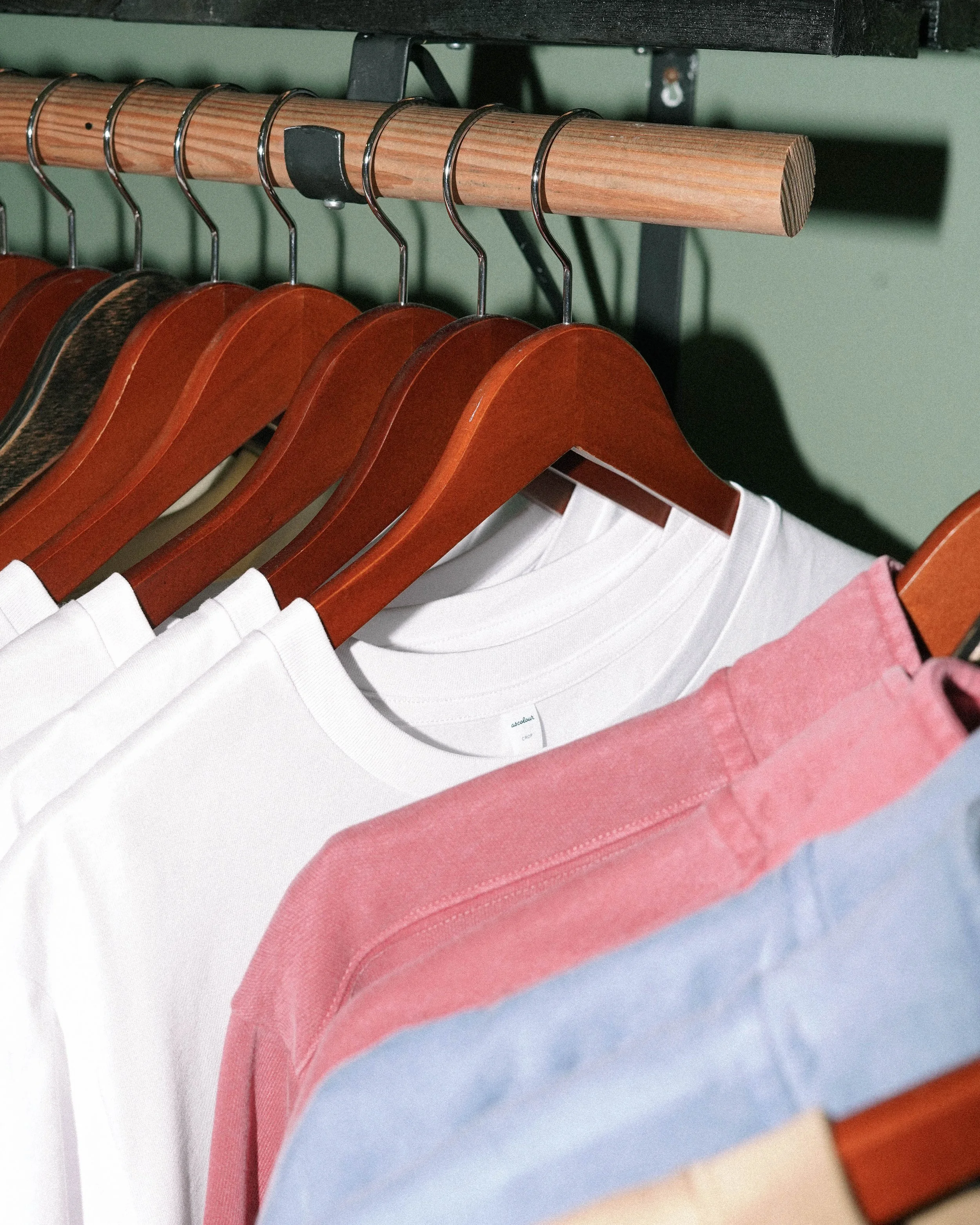Clothes hanging on wooden hangers in a closet, including white t-shirts, pink and blue tops.